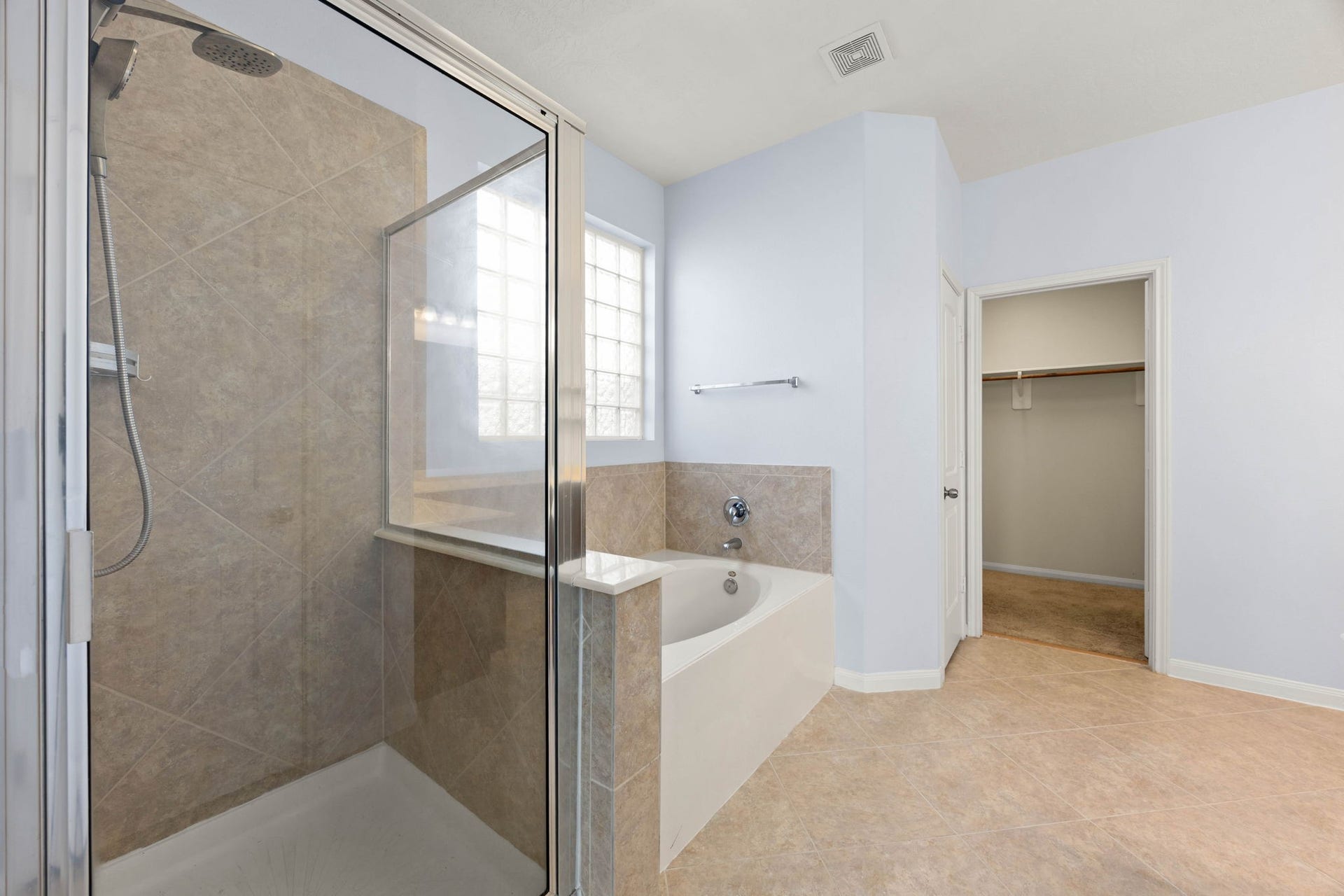 A luxurious spa-like retreat awaits in the primary en-suite, featuring a separate deep soaking tub and a dedicated glass-enclosed walk-in shower. The spaces are elegantly tied together with neutral diagonal tile surrounds, a privacy-enhancing glass block window that lets in soft natural light, and direct access to a spacious walk-in closet just steps away.