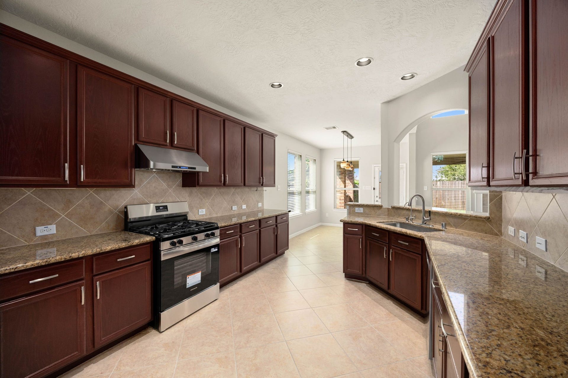 This reverse angle of the spacious kitchen highlights the impressive length of the granite countertops and the extensive cabinetry layout. The view emphasizes the integrated flow from the cooking zonefeaturing a stainless steel gas range and matching vent hoodstraight into the sunlit breakfast nook and out toward the backyard, making it an ideal setup for hosting and daily meal prep.