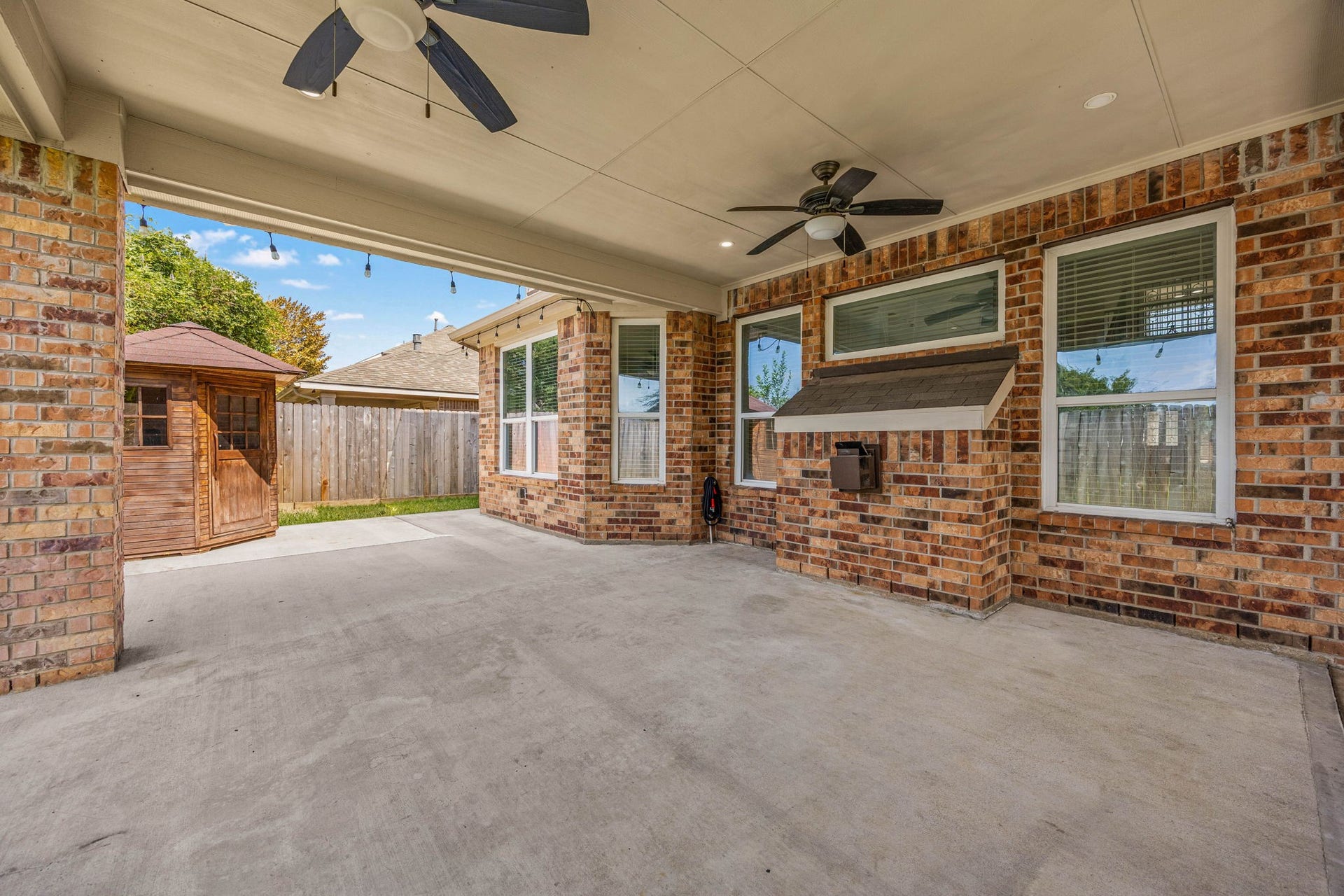 This reverse angle of the expansive covered patio emphasizes the generous width of the concrete slab and the continuous brick craftsmanship that ties the outdoor living space to the main home. The functional design includes dual ceiling fans, a unique built-in brick service station with a shingled awning, and a clear view out to the fully fenced yard featuring a premier standalone wooden barrel-style sauna that offers a private, spa-like wellness retreat right at home.