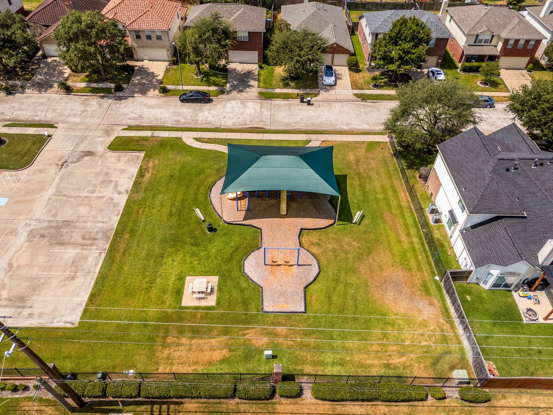 A dedicated neighborhood play area sits adjacent to the community pool complex, offering an ideal spot for outdoor play and gatherings. It features a swing set and a multi-play structure fully protected by a large green canvas canopy for essential sun protection, alongside a concrete parking lot and a shaded picnic table for comfortable family outings.