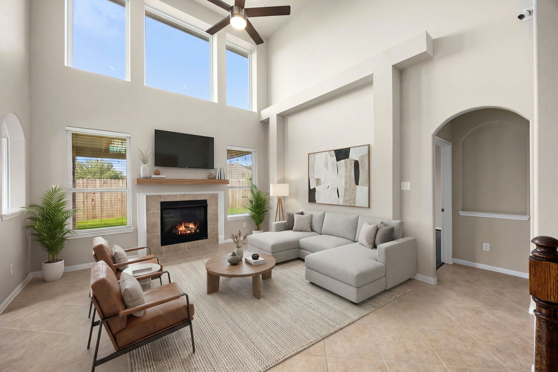 Soaring two-story ceilings and an impressive wall of upper windows define this spectacular living room, filling the space with an exceptional amount of natural light. A cozy fireplace with a custom wood mantel serves as the room's focal point, beautifully complemented by neutral tile flooring, a modern ceiling fan, and elegant architectural arches that transition seamlessly into adjacent living spaces. *Virtually Staged