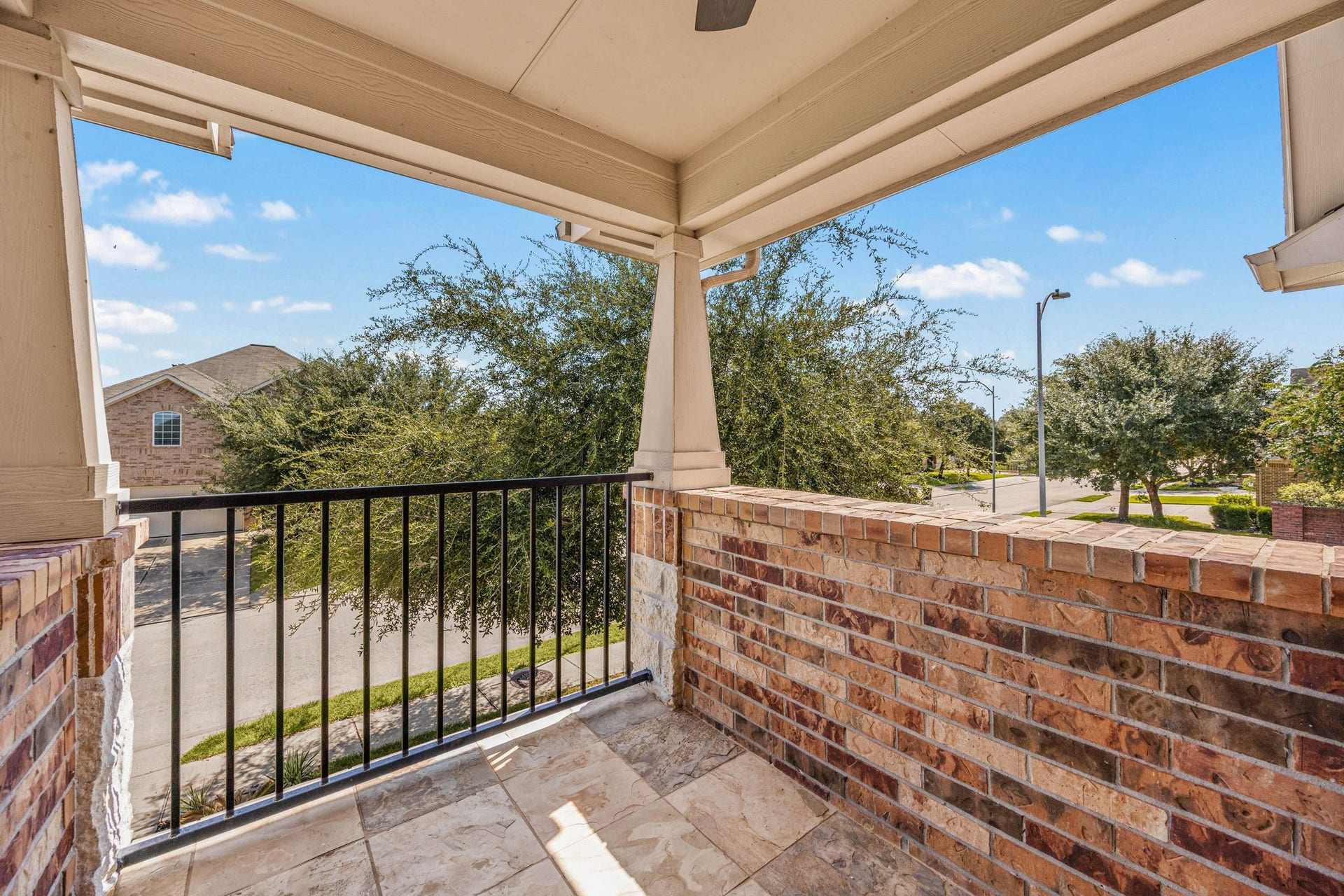 Step outside onto this private covered balcony, a unique second-story retreat offering a peaceful vantage point over the quiet neighborhood street. The space is beautifully finished with durable tile flooring, sturdy brick accents, a classic wrought-iron railing, and a covered ceiling with a cooling fan, making it the perfect spot for morning coffee or evening relaxation among the treetops.