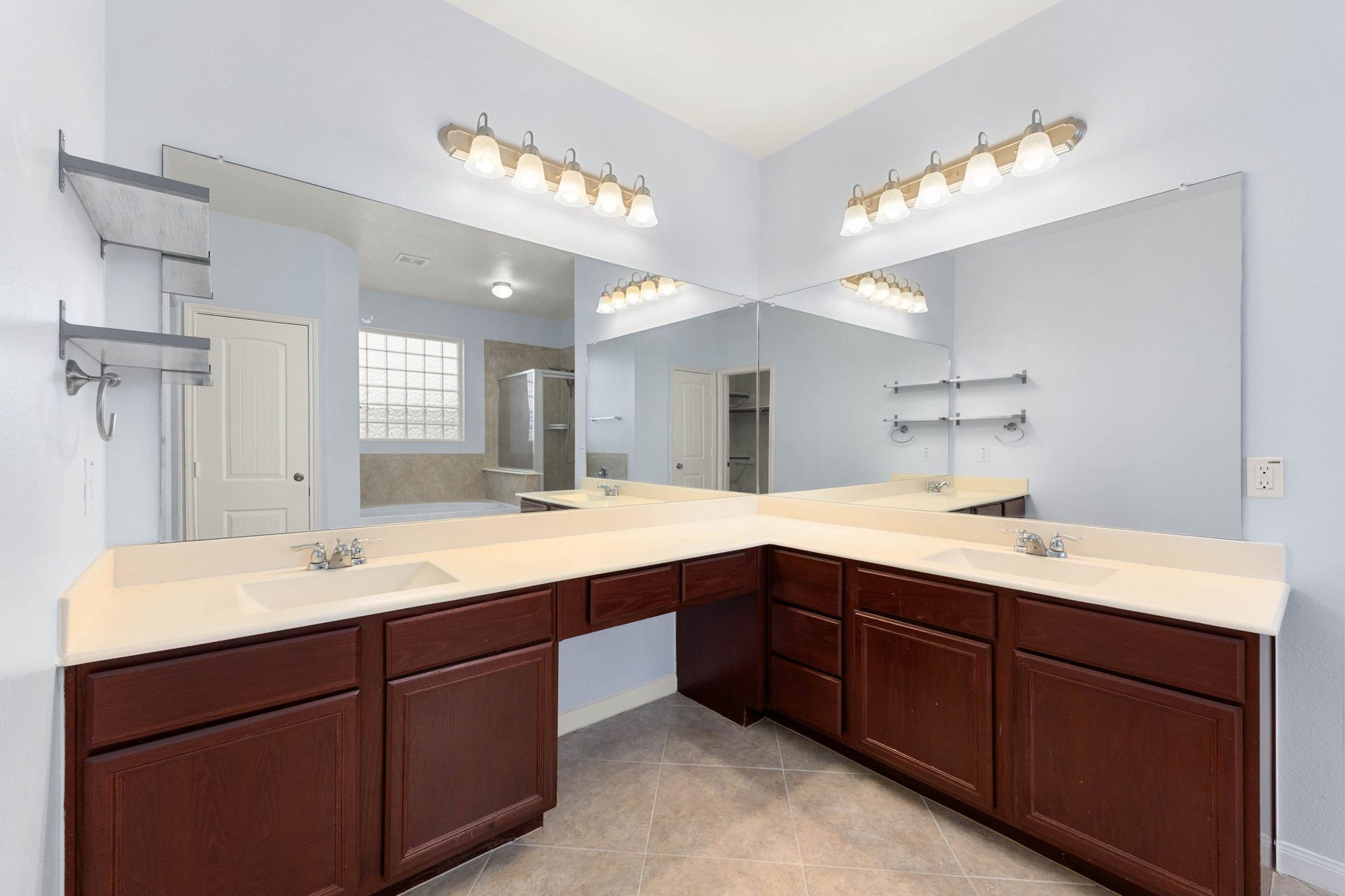 An expansive wrap-around vanity anchors this primary en-suite bathroom, offering an abundance of counter space and dedicated dual-sink stations. The functional L-shaped layout features rich dark cabinetry with integrated storage, a built-in makeup vanity knee space, matching multi-bulb vanity light fixtures, and massive wrap-around mirrors that amplify the room's open, airy feel.