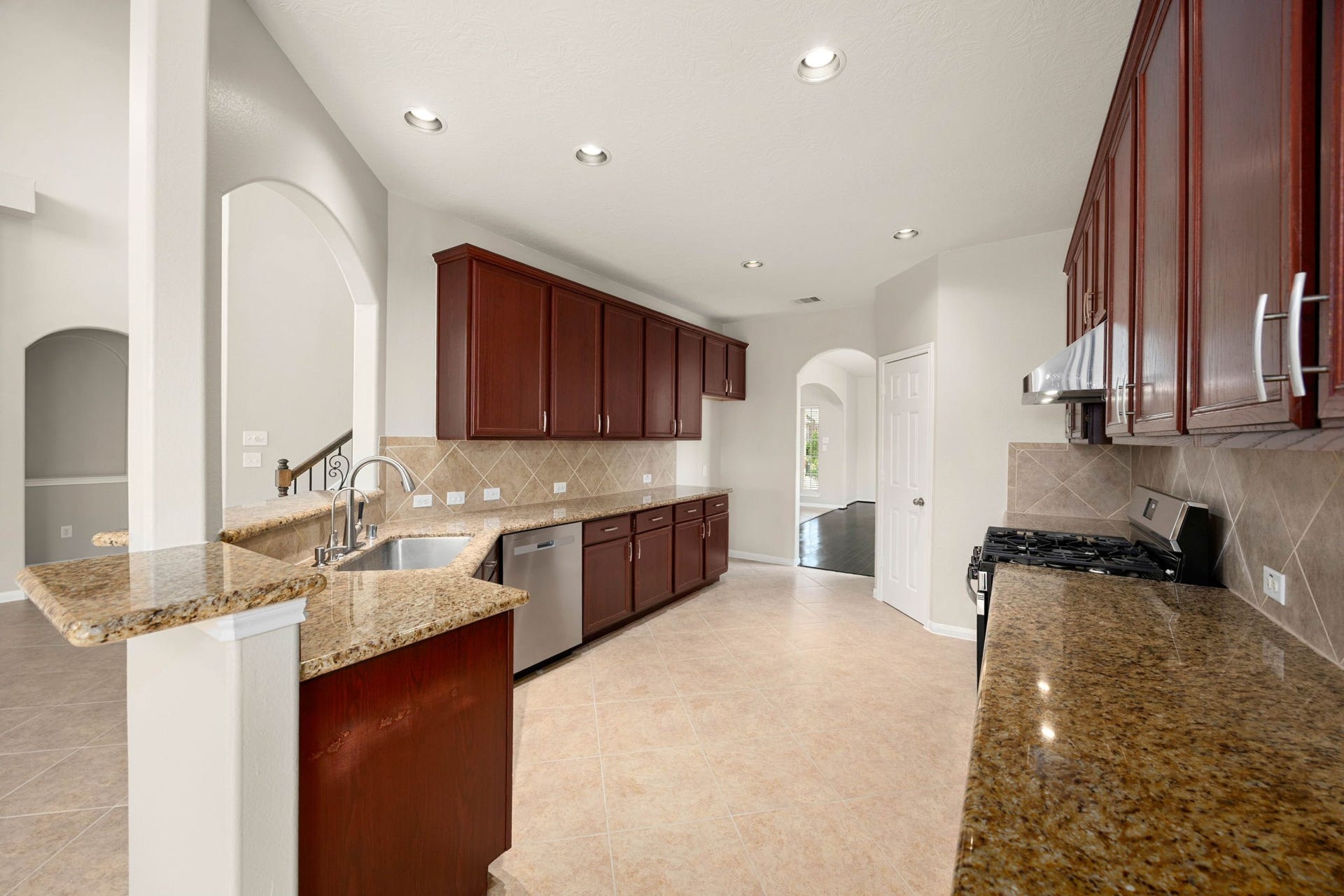 Abundant granite counter space and rich, dark-stained cabinetry define this spacious kitchen, designed for maximum efficiency and storage. The functional layout features sleek stainless-steel appliances, including a under-mount double sink with a modern high-arc faucet, a gas range, and a diagonal tile backsplash, all illuminated by recessed lighting.