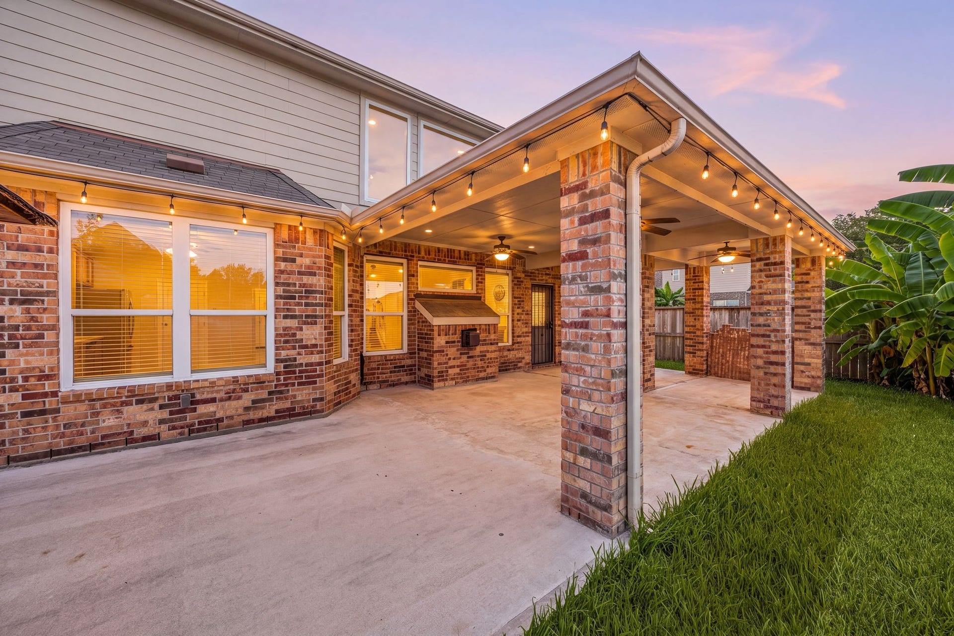 An expansive covered patio addition transforms this backyard into an outdoor entertainer's paradise, highlighted by robust brick pillars. The massive concrete extension features multiple integrated ceiling fans, a functional built-in service pass-through structure, and a deep overhang that provides continuous shade and comfortable outdoor living space year-round.