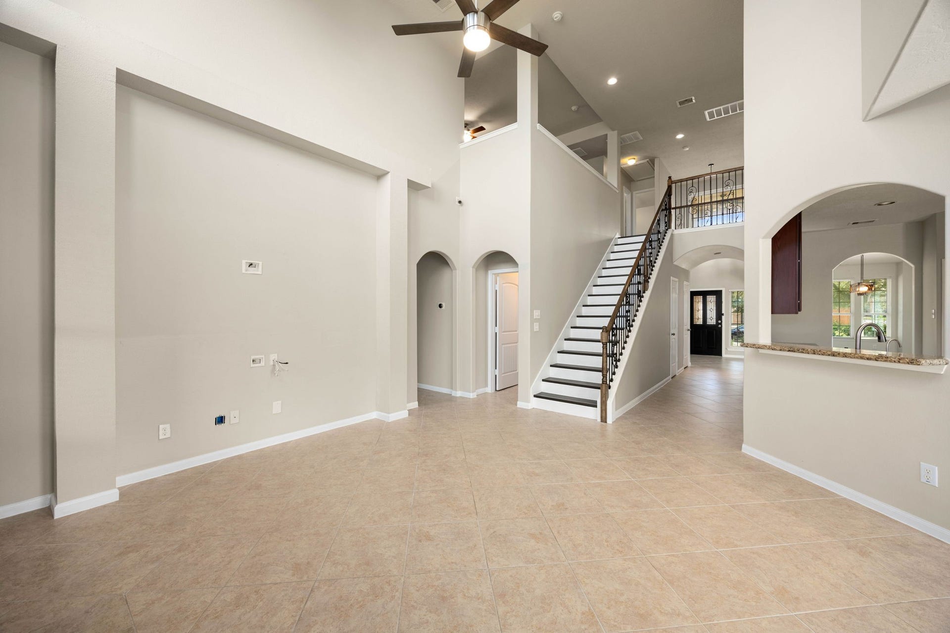 This commanding perspective from the family room showcases the home's impressive vertical scale, drawing your eye up to the high ceilings and the sweeping staircase accented by elegant wrought-iron spindles. The neutral tile flooring stretches past the arched entryways and a half-wall kitchen cutout toward the bright foyer and front door, while the upper-level catwalk overlooks this grand central living space.
