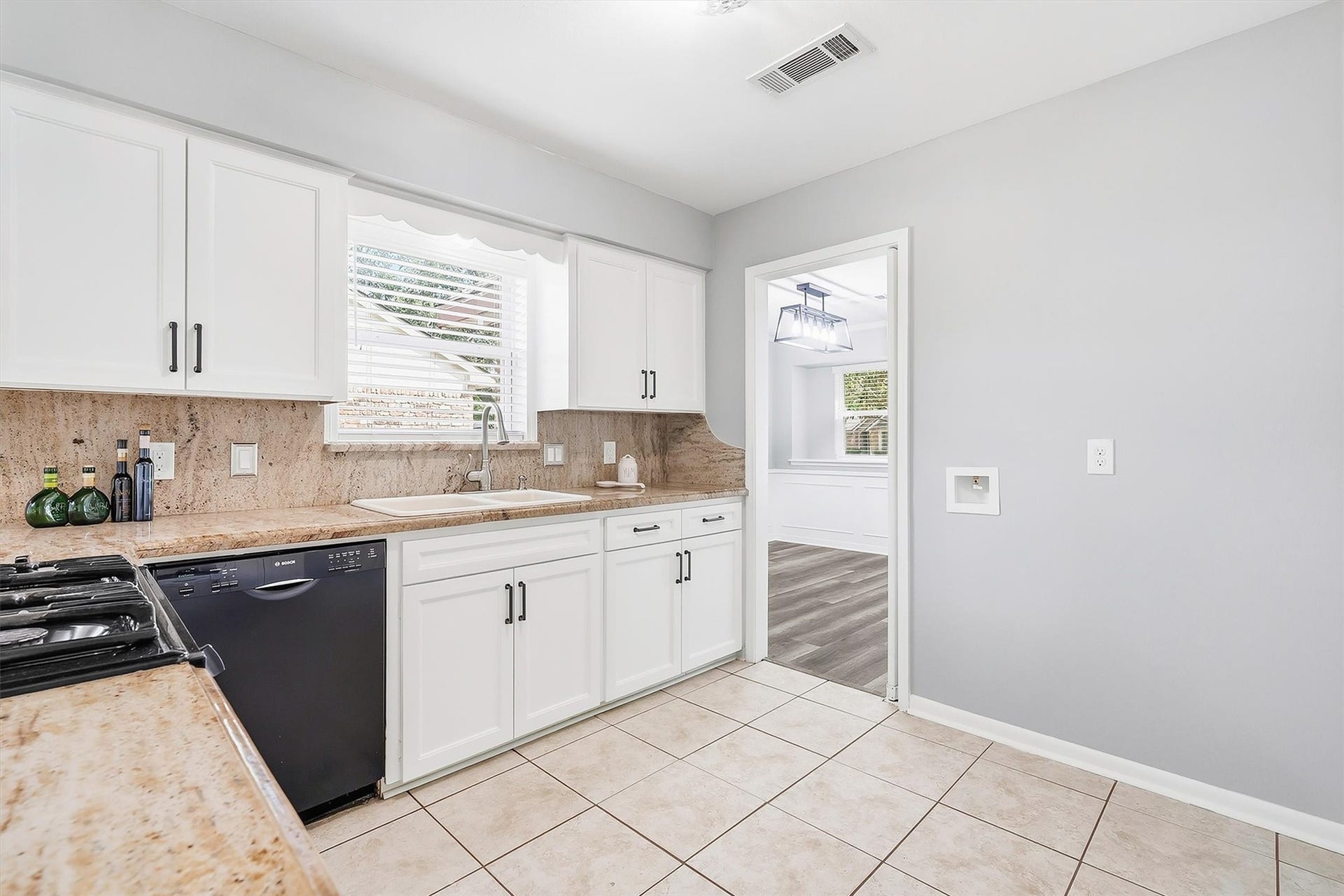 2503 Moss Hill Drive, Houston, TX 77080 Bright kitchen with sleek white cabinetry, granite countertops, and a modern black dishwasher. A window above the sink offers natural light, and the adjacent room features elegant wainscoting and a stylish light fixture.