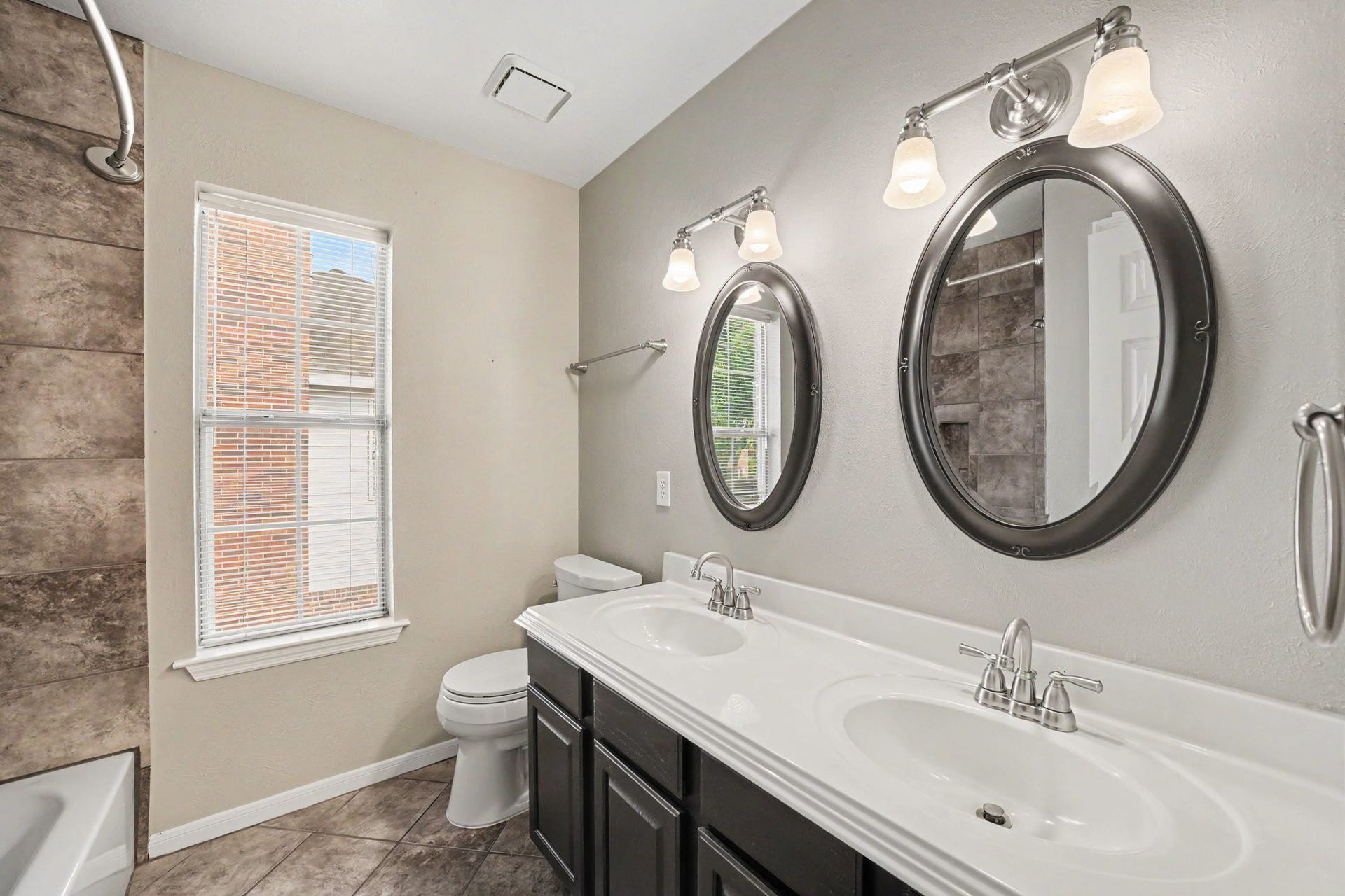 17510 Tall Maple Court, Houston, TX 77095 The upstairs secondary bathroom features a double-sink vanity with contrasting dark cabinetry, individual framed mirrors, and updated lighting. A tub and shower combination with floor-to-ceiling tile surround completes the space, while a large window brings in natural light.