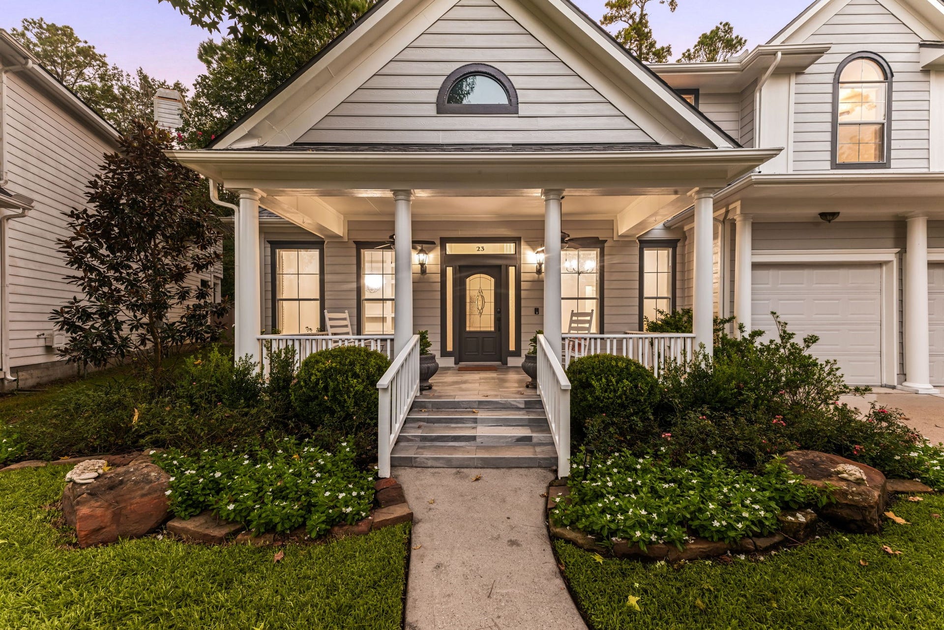 23 Pipers Meadow Street, The Woodlands, TX 77382 Inviting front entrance featuring an expansive covered porch, beautiful symmetry, soft exterior lighting, and lush low-maintenance landscaping. Perfect for enjoying quiet evenings in The Woodlands.