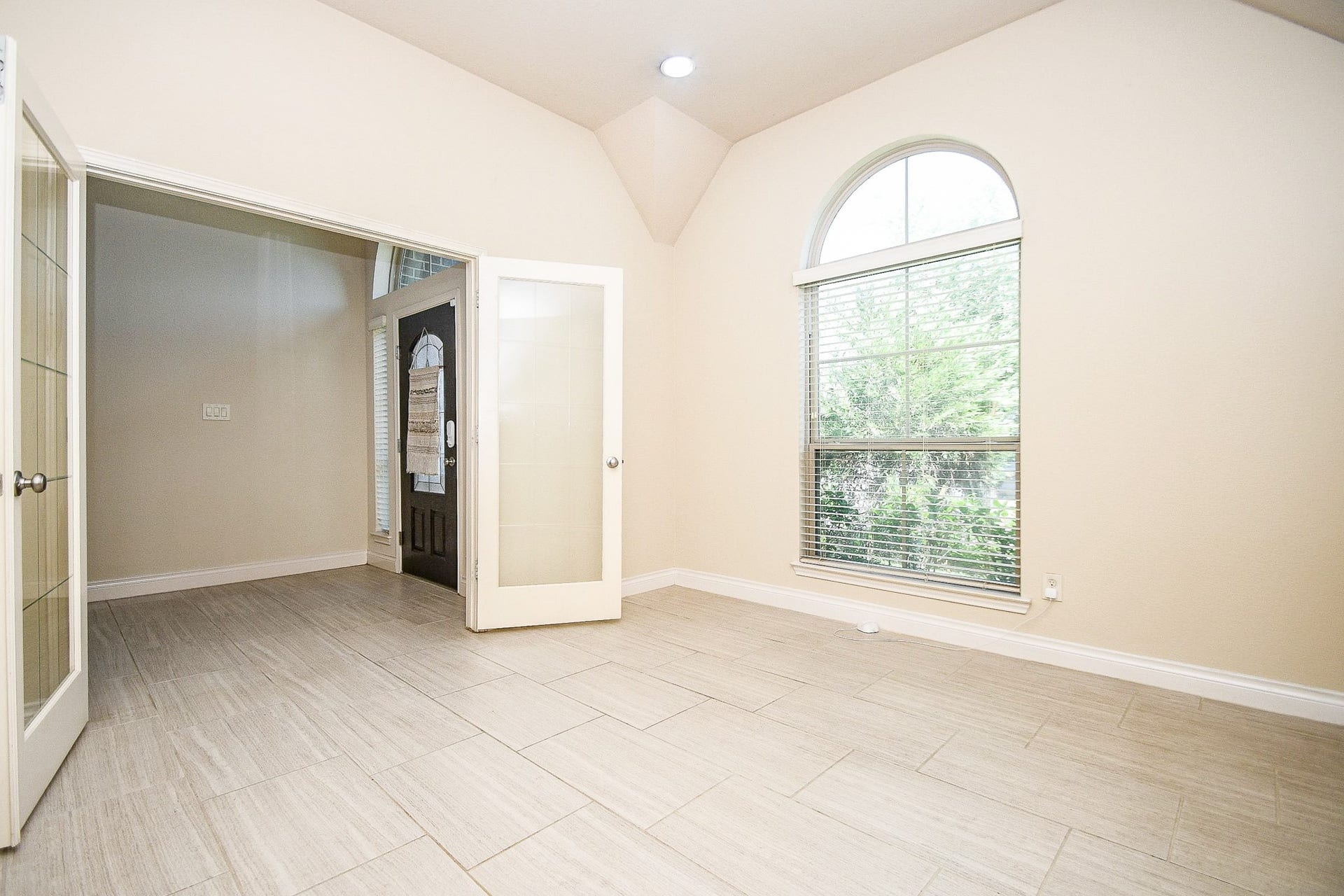 A bright and welcoming entryway boasts large windows, a vaulted ceiling, and neutral tile flooring, complemented by elegant glass double doors to the study, creating an inviting atmosphere filled with natural light.