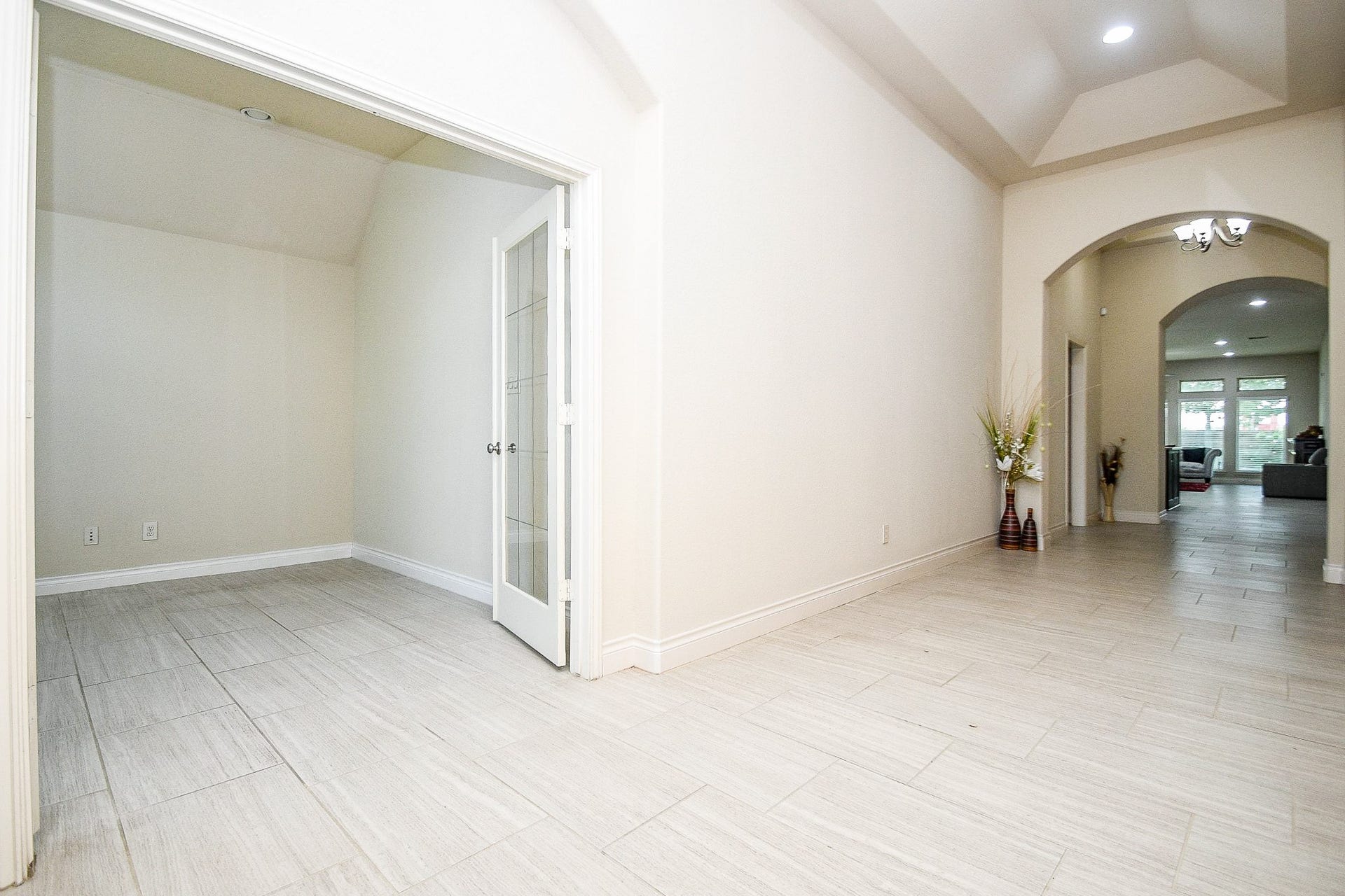A spacious, bright hallway with light tiles and neutral walls leads to a room with elegant double glass doors, opening into a modern, inviting living area filled with natural light from large windows, perfect for comfortable living.