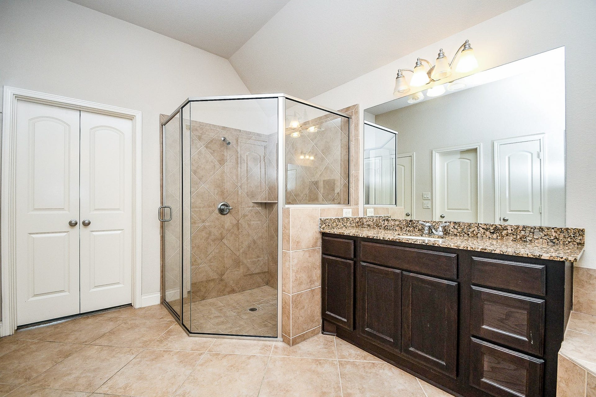 This spacious bathroom features a large glass-enclosed shower with beige tile, a double-sink vanity with dark wood cabinets and granite countertops, and ample lighting. The neutral tones create a clean and inviting atmosphere.