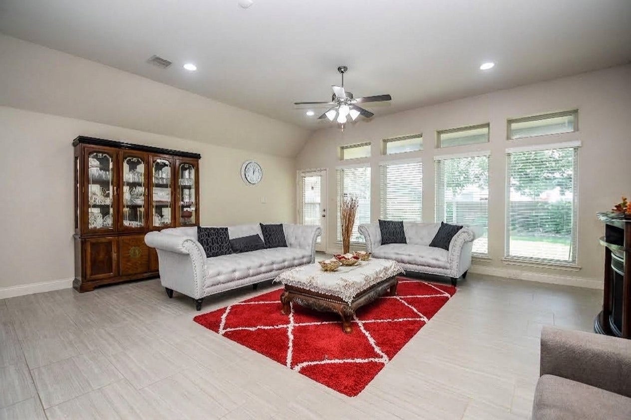 Spacious living room with abundant natural light, featuring elegant seating, a stylish red area rug, and classic wooden cabinetry.