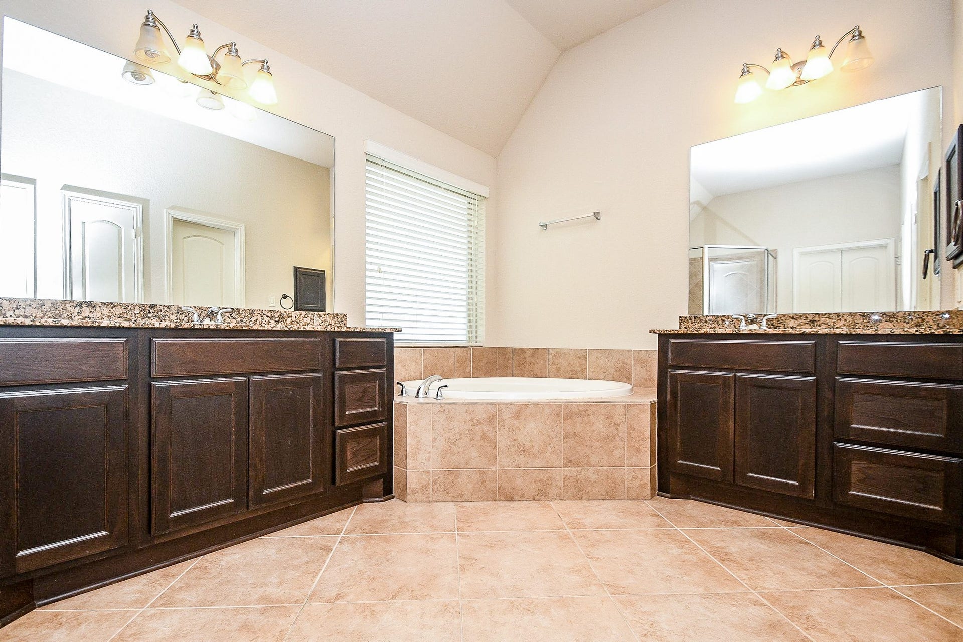 Discover a luxurious bathroom with dual vanities and granite countertops, complemented by elegant dark wood cabinetry and a central soaking tub, all illuminated by bright lighting and a large window that enhances the spacious, tiled ambiance.