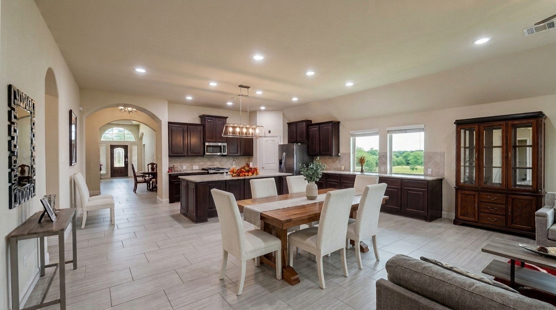 Spacious open-concept living area featuring a modern kitchen with dark wood cabinetry, stainless steel appliances, and a large island. The dining and living areas are bright and airy with ample natural light from large windows overlooking serene greenery.  AI designed