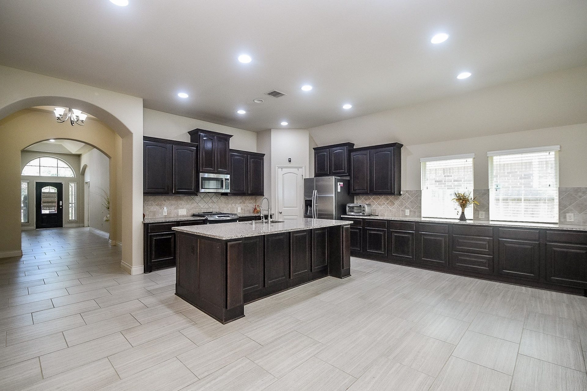 Discover a spacious kitchen with elegant dark wood cabinetry, a large central island with granite countertops, and stainless steel appliances, all illuminated by natural light from multiple windows, seamlessly flowing into a welcoming tiled hallway.