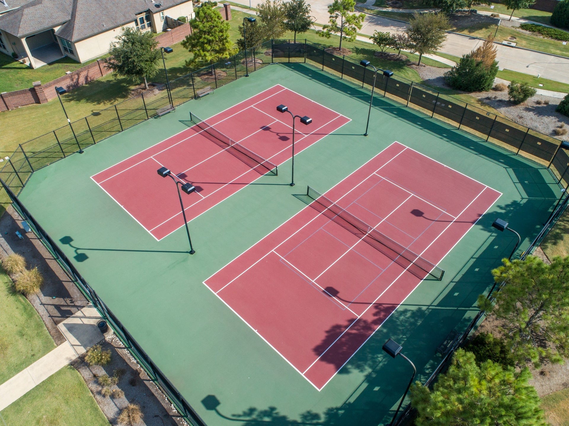 This photo shows a well-maintained tennis court facility with two courts, surrounded by a fence and greenery. Ideal for recreational activities, it offers ample space and lighting for evening play.