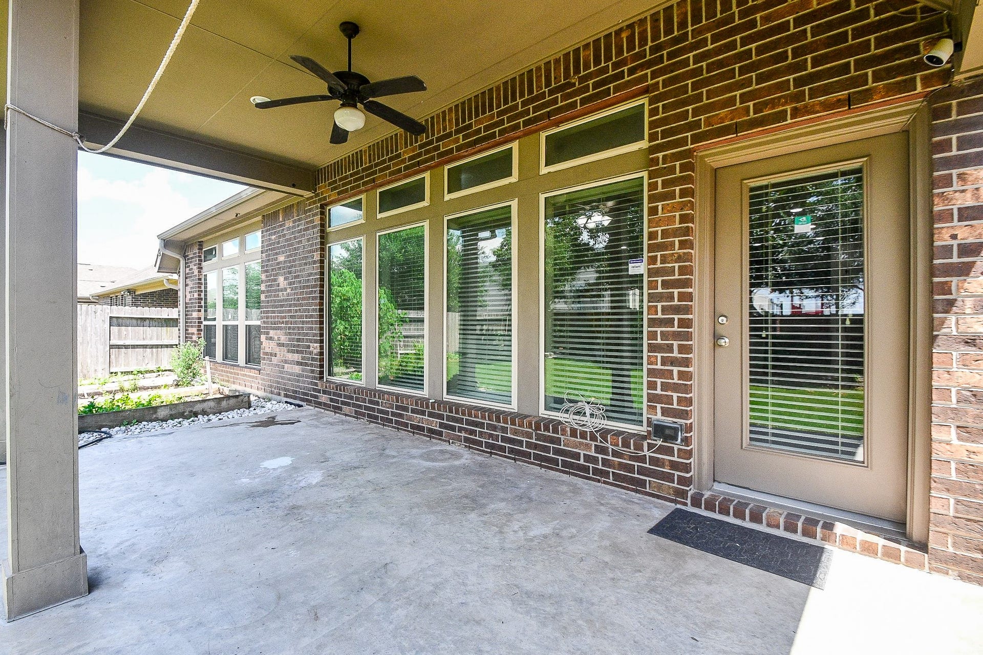 Experience effortless outdoor living on a spacious covered patio, featuring a ceiling fan, elegant brick exterior, and large windows that frame a beautifully maintained backyard, ideal for relaxation and entertaining.