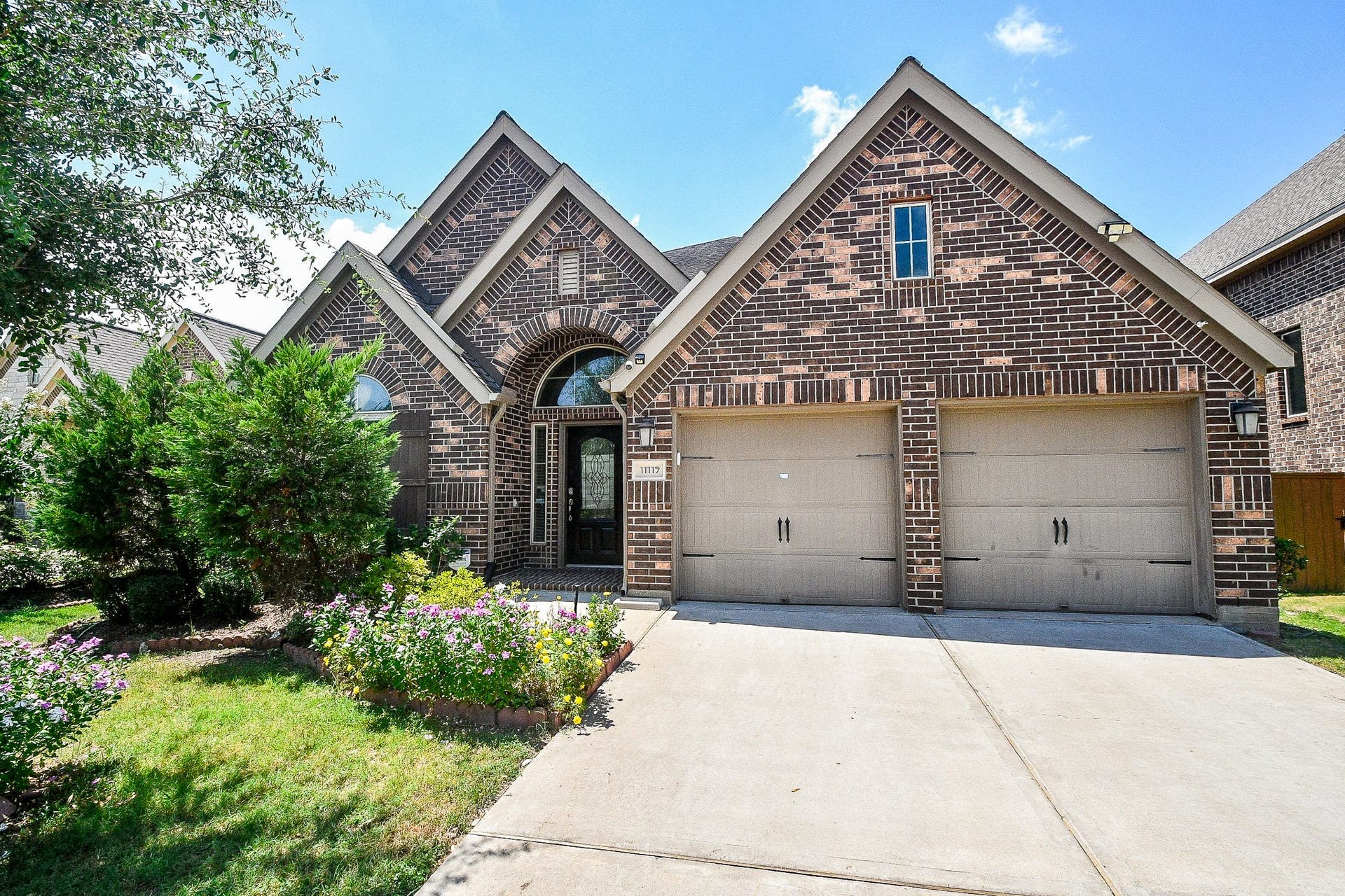 Charming brick home with a two-car garage and a welcoming arched entryway, nestled amidst lush greenery and vibrant flowers for irresistible curb appeal.
