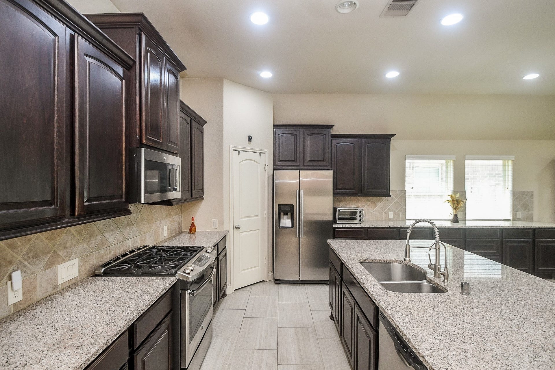Experience modern elegance in this kitchen with dark wood cabinetry, sleek granite countertops, stainless steel appliances, and a spacious island featuring dual sinks, all beautifully illuminated by recessed lighting for a bright, open feel.