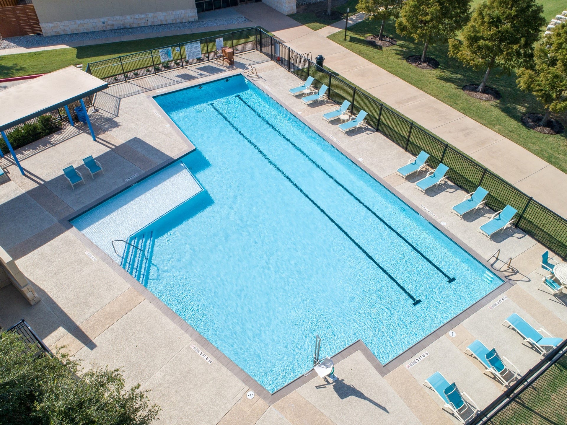 This photo showcases a large, inviting community swimming pool with clear water, surrounded by a spacious deck with several lounge chairs and a shaded seating area. It's set in a well-maintained outdoor space with adjacent greenery, perfect for relaxation and recreation.