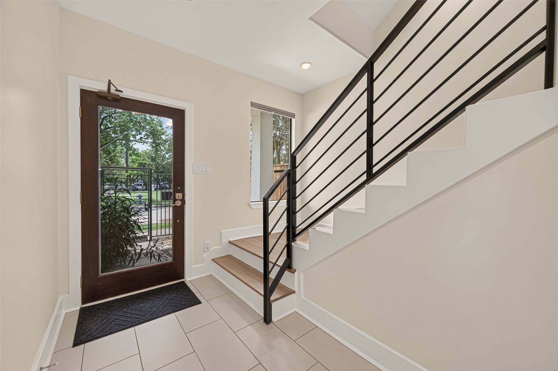 Bright entryway with a modern glass door, tile flooring, and a sleek staircase. Large window offers natural light and a view of the greenery outside.