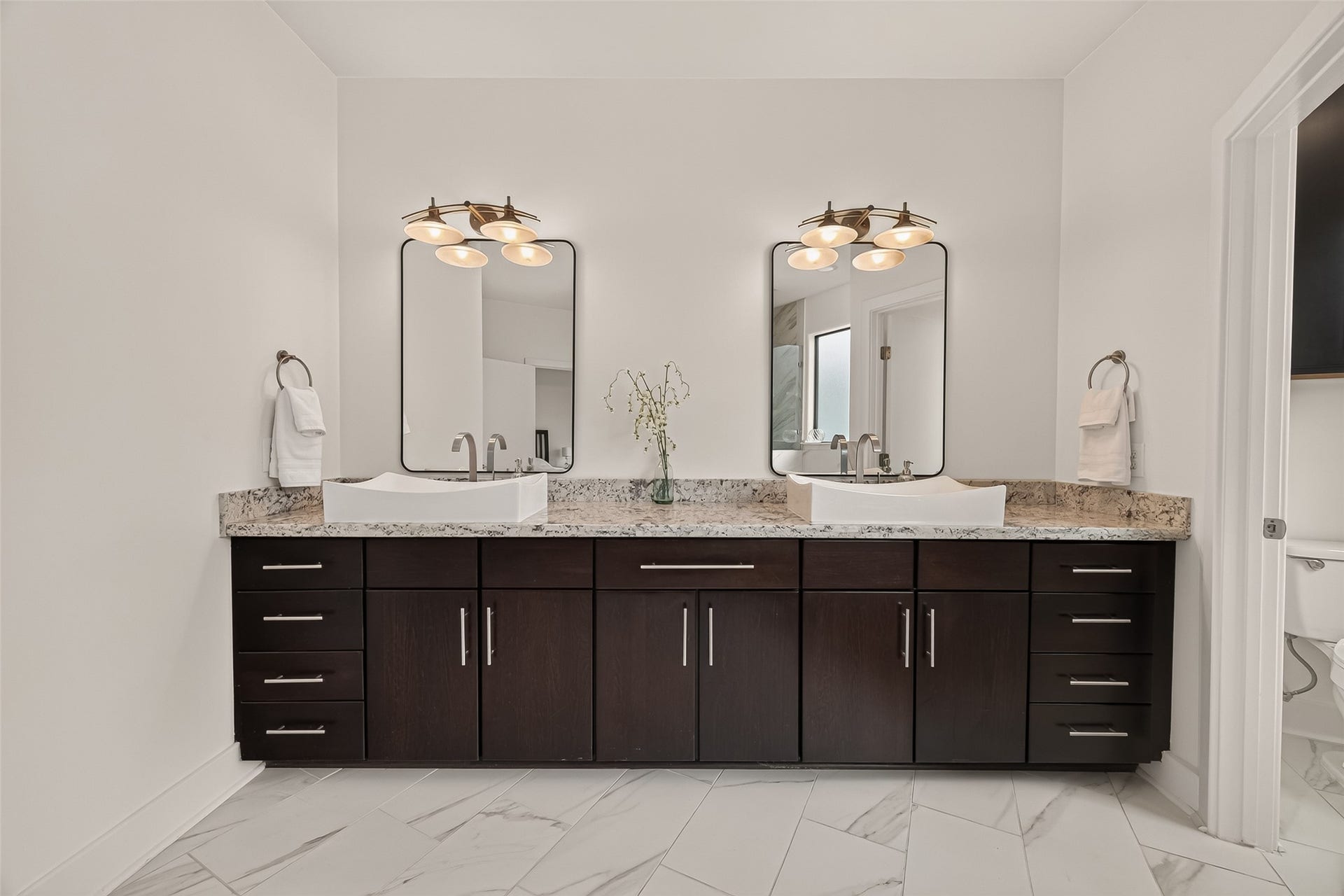 The primary bathroom features a modern double vanity with dark wood cabinets, granite countertops, and elegant vessel sinks. It includes large mirrors and stylish lighting, offering a clean and contemporary feel.