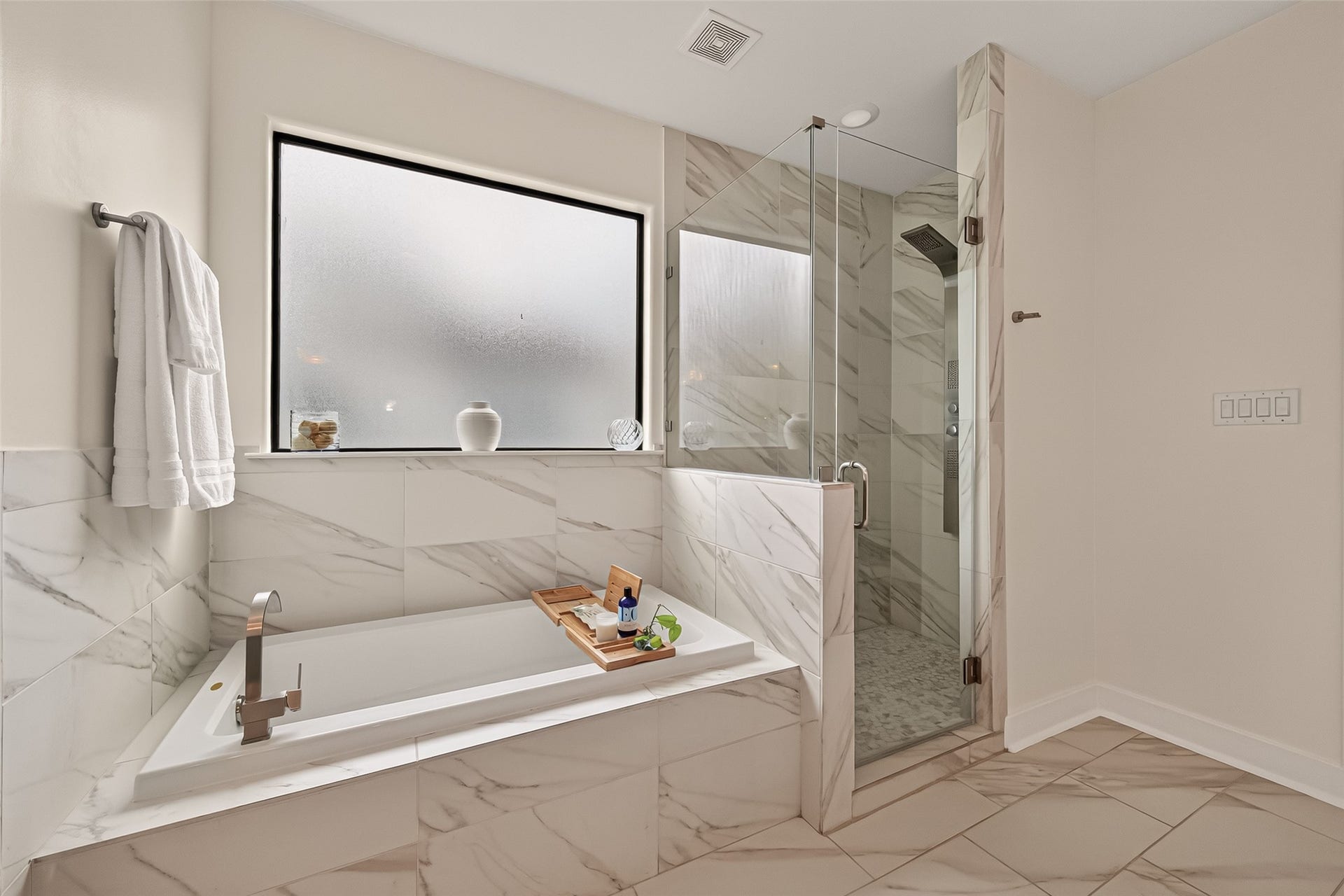 The primary bathroom also features a large soaking tub with elegant marble tile, a sleek glass-enclosed shower, and a frosted window for privacy.