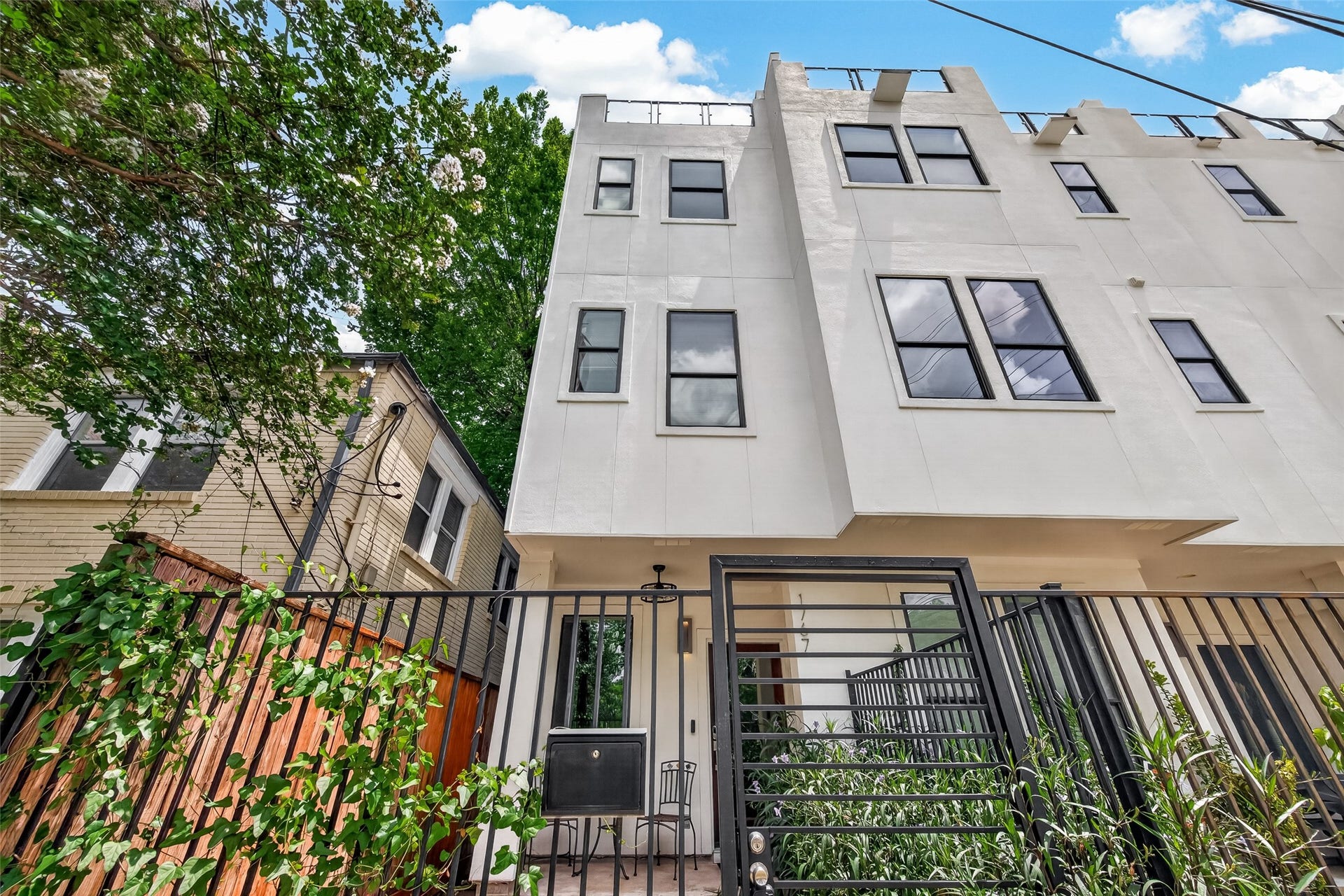 Modern three-story townhouse . Features a gated entrance and a small patio area, surrounded by greenery. Ideal for those seeking contemporary urban living.