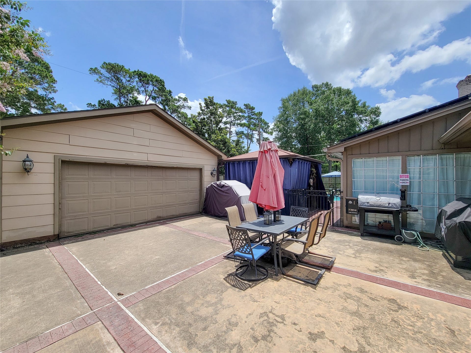 6235 Rolling Water Drive, Houston, TX 77069 A spacious backyard patio area with a dining set and umbrella, a grill, and ample space for outdoor activities. It is bordered by a garage and features greenery in the background, offering a private and inviting space for entertaining.