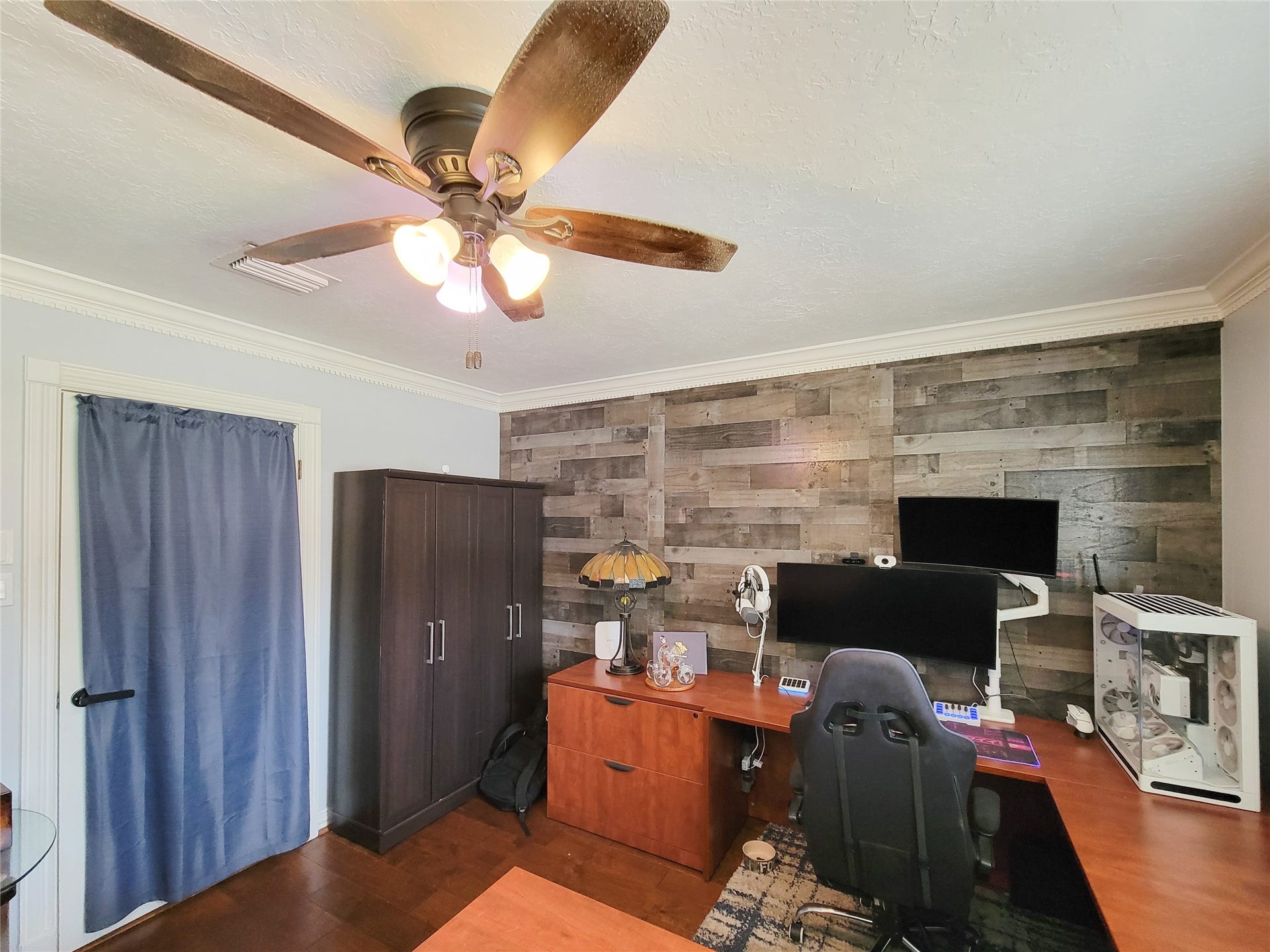 6235 Rolling Water Drive, Houston, TX 77069 A cozy home office featuring a modern ceiling fan, wooden accent wall, and ample workspace with a large desk and shelving. The room includes a comfortable office chair, built-in computer setup, and stylish lighting, creating an inviting and functional work environment.