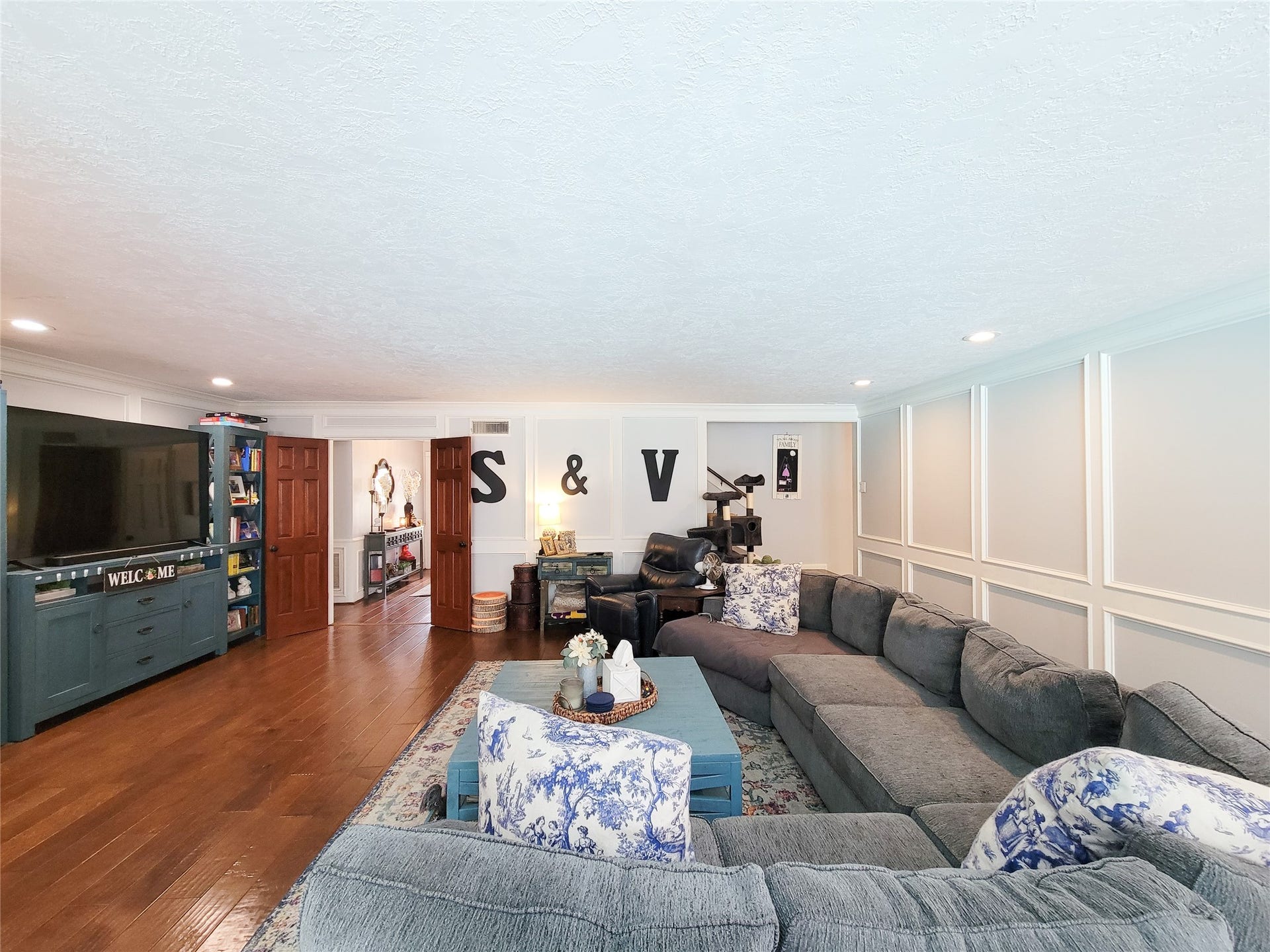 6235 Rolling Water Drive, Houston, TX 77069 Spacious living room with elegant wood flooring, a large sectional sofa, and ample natural light. Features a cozy TV area, decorative wall paneling, and open entryways leading to adjacent rooms.