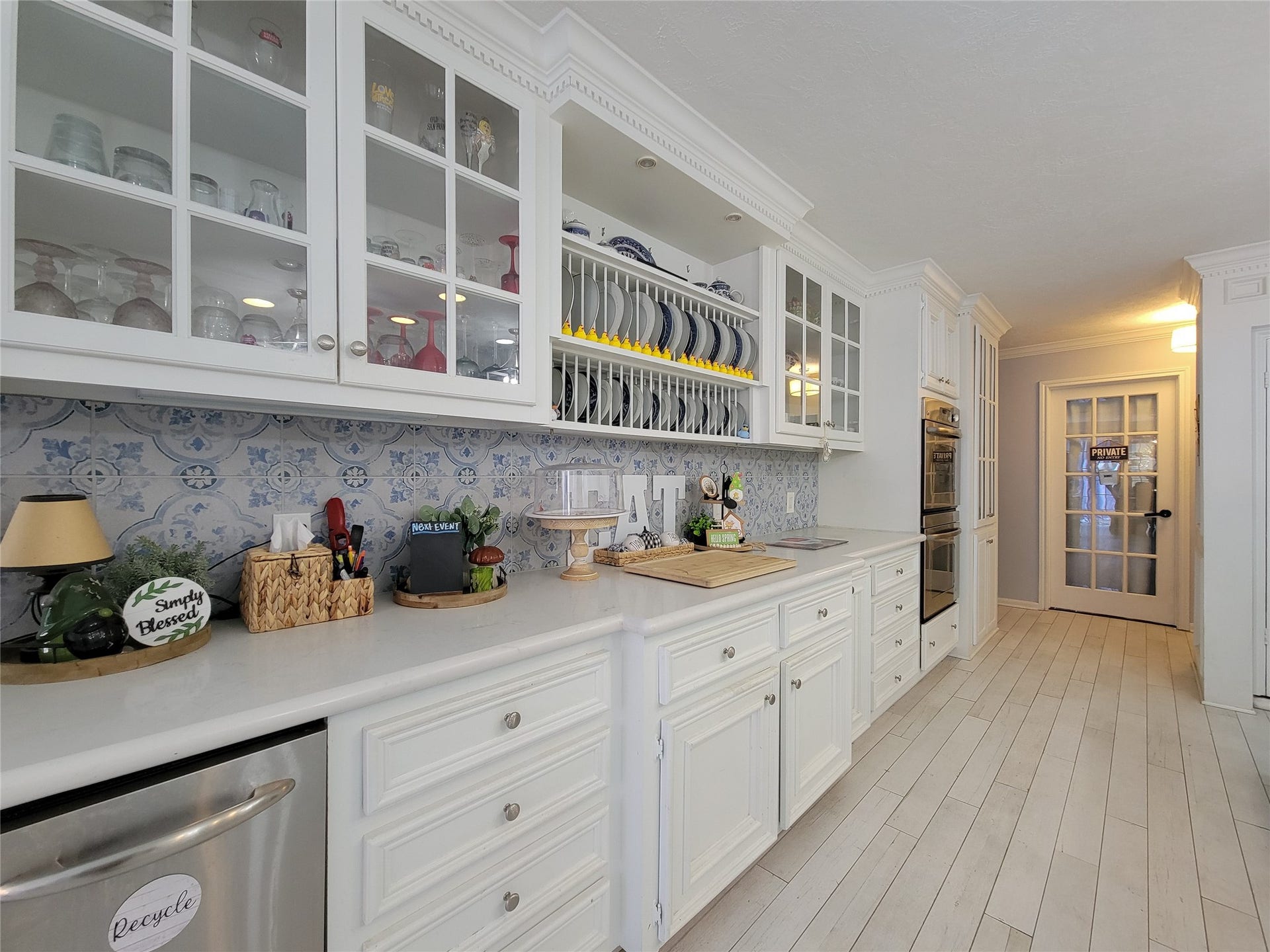 6235 Rolling Water Drive, Houston, TX 77069 This kitchen features elegant white cabinetry with glass doors, a decorative blue and white tile backsplash, and ample counter space. It includes a built-in oven and a dishwasher, with a bright, inviting atmosphere enhanced by light wood flooring and recessed lighting.
