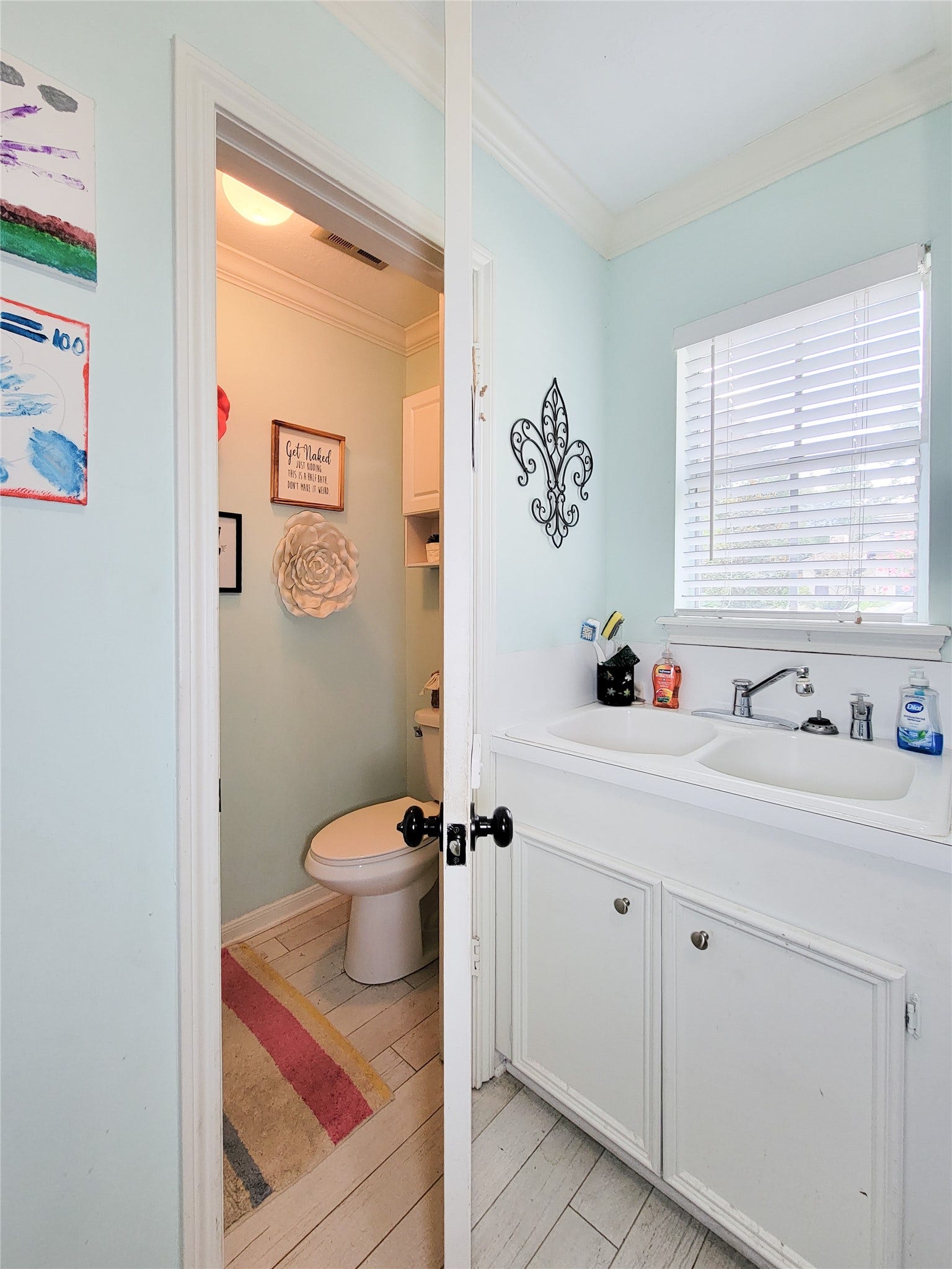 6235 Rolling Water Drive, Houston, TX 77069 This photo showcases a compact bathroom with pastel blue walls, featuring a toilet, a white sink with cabinets, and a window with blinds. Decorative elements include wall art and a small rug, creating a cozy, inviting space.