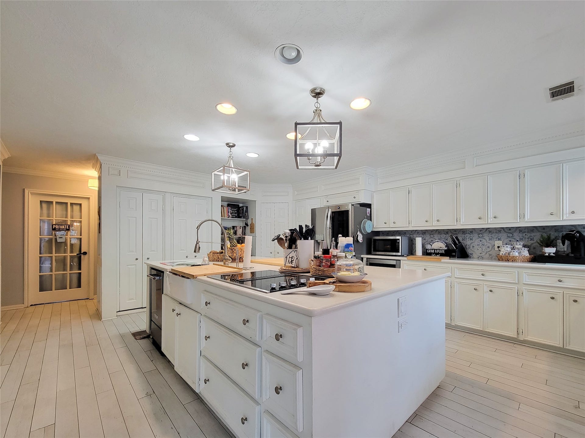 6235 Rolling Water Drive, Houston, TX 77069 This spacious kitchen features modern lighting, ample white cabinetry, and a large island with a cooktop and sink. The light wood flooring and stainless steel appliances add a contemporary touch, while a glass-paneled door leads to additional storage space.