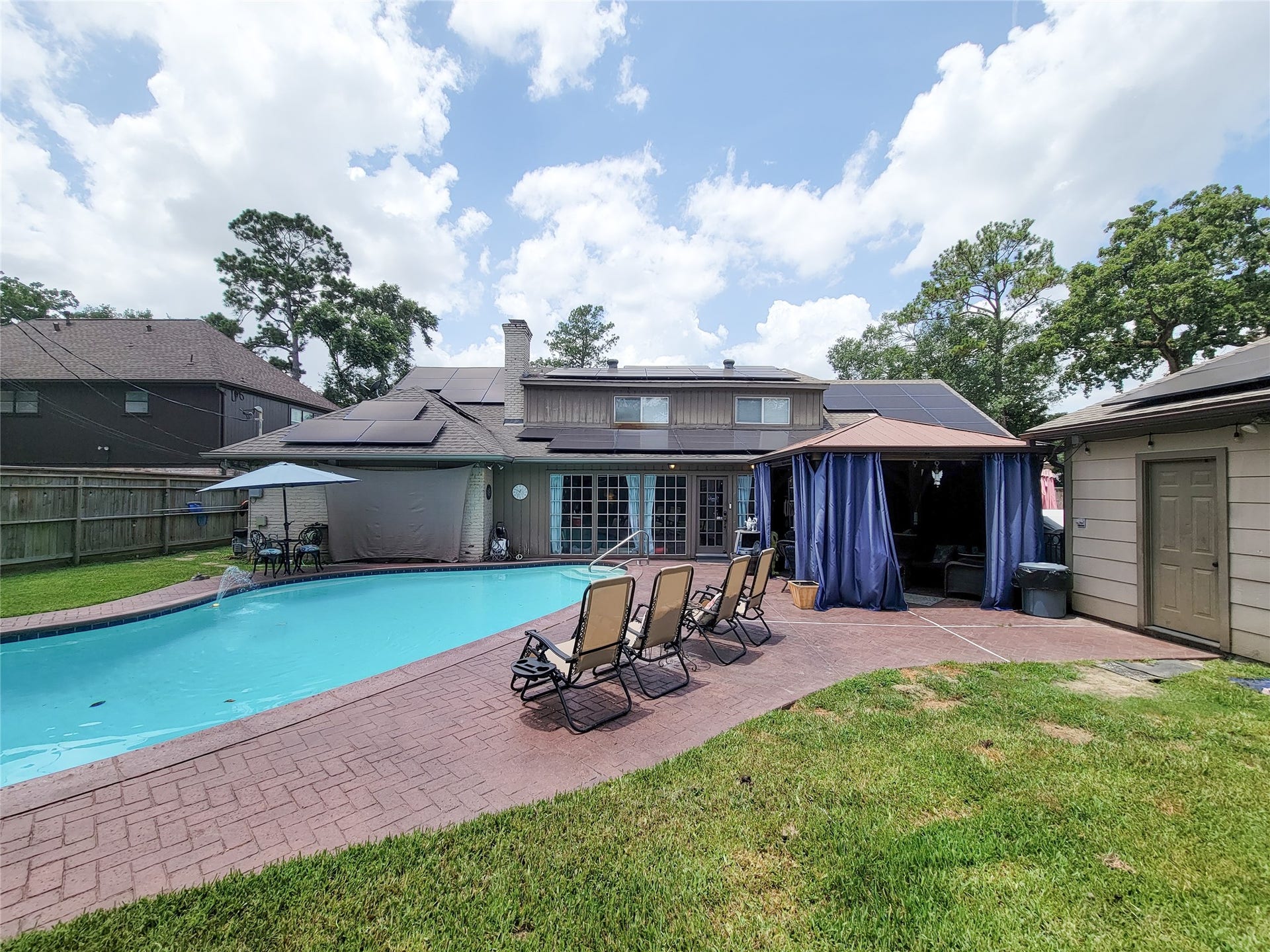 6235 Rolling Water Drive, Houston, TX 77069 This photo features a spacious backyard with a large swimming pool, surrounded by a brick patio. There's ample seating with several reclining chairs, a covered outdoor area with curtains, and mature trees providing natural shade. The house has solar panels and a fence for privacy, offering a perfect blend of relaxation and functionality.