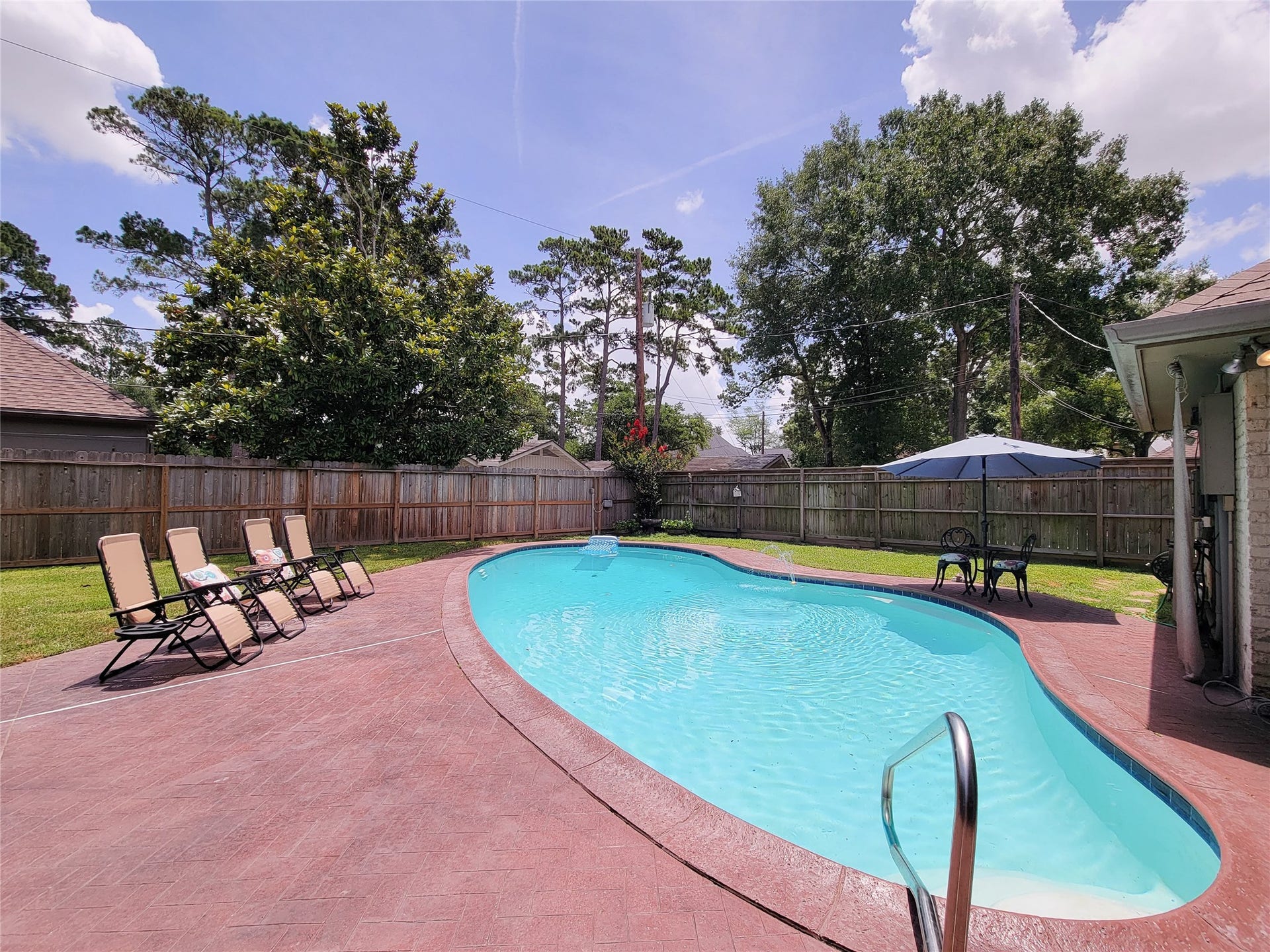 6235 Rolling Water Drive, Houston, TX 77069 This photo showcases a spacious backyard with a curvy pool, surrounded by a well-maintained patio. There are several lounge chairs and a seating area with an umbrella, perfect for relaxation and entertaining against a backdrop of mature trees and a wooden fence.