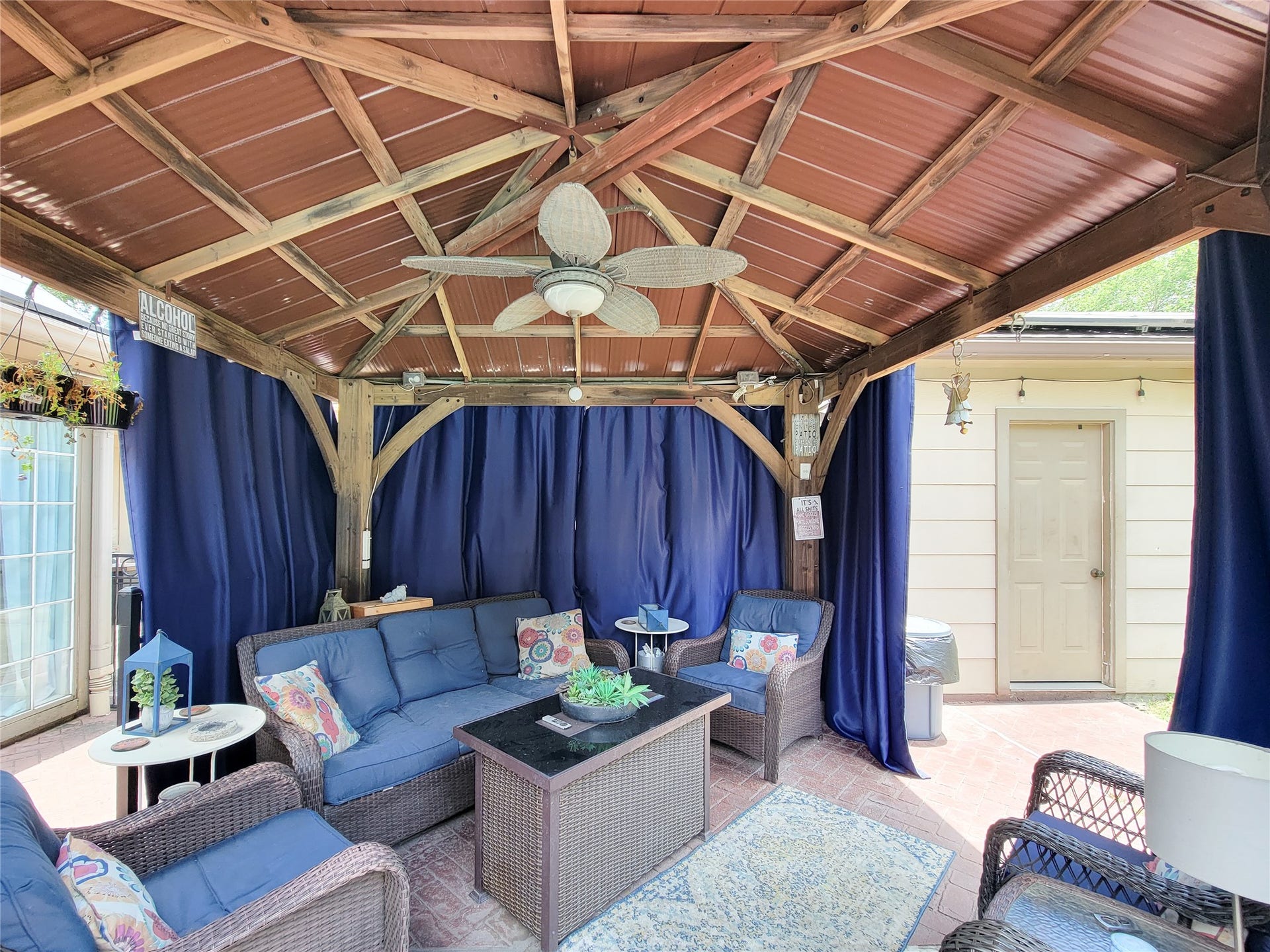 6235 Rolling Water Drive, Houston, TX 77069 Cozy outdoor patio with a wooden gazebo, featuring comfortable wicker seating, a ceiling fan, and blue curtains for privacy. It's an inviting space for relaxation and entertaining.