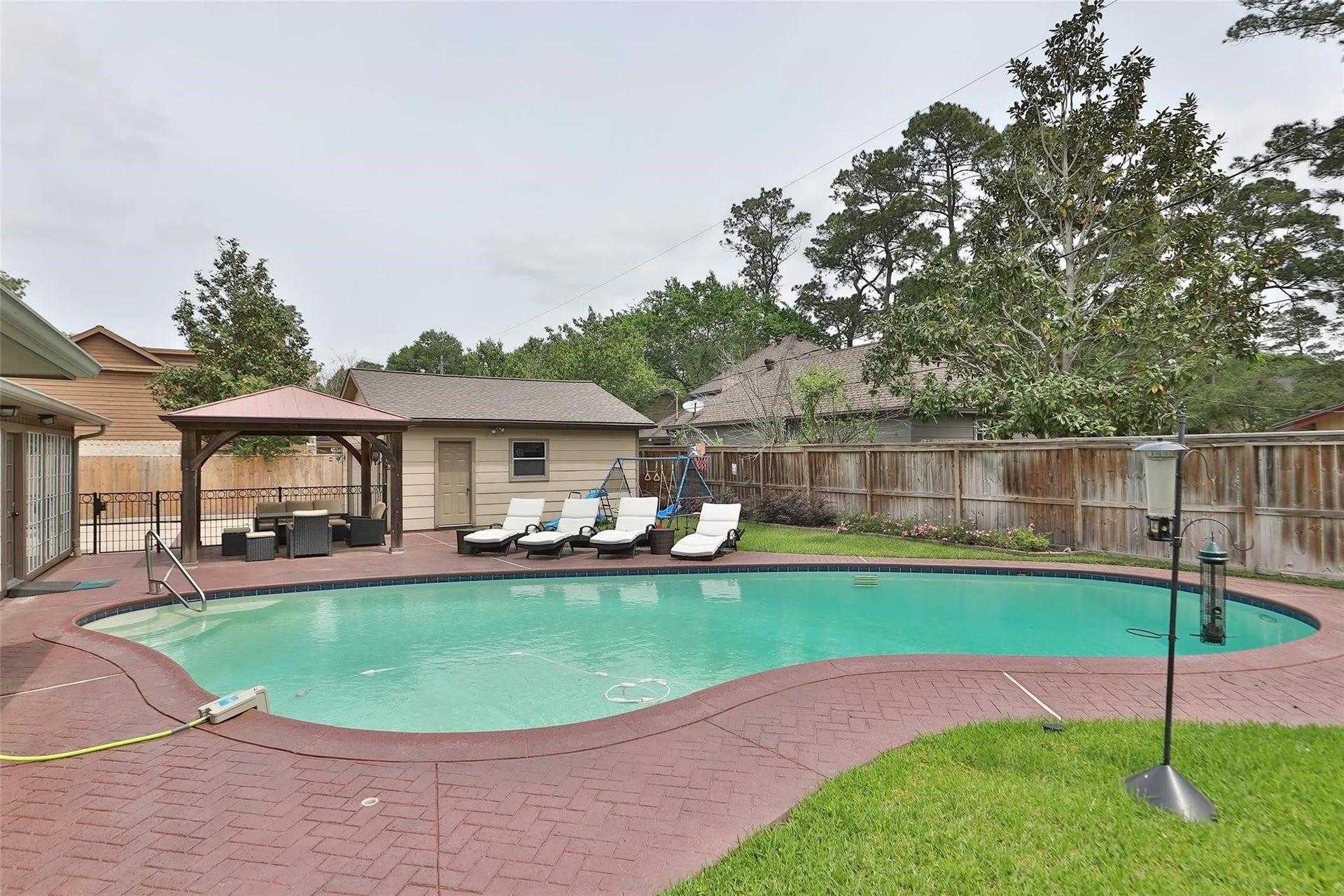 6235 Rolling Water Drive, Houston, TX 77069 This backyard features a spacious, curved swimming pool surrounded by a brick patio. The area is enclosed by a wooden fence and bordered by mature trees, offering privacy and a serene atmosphere.