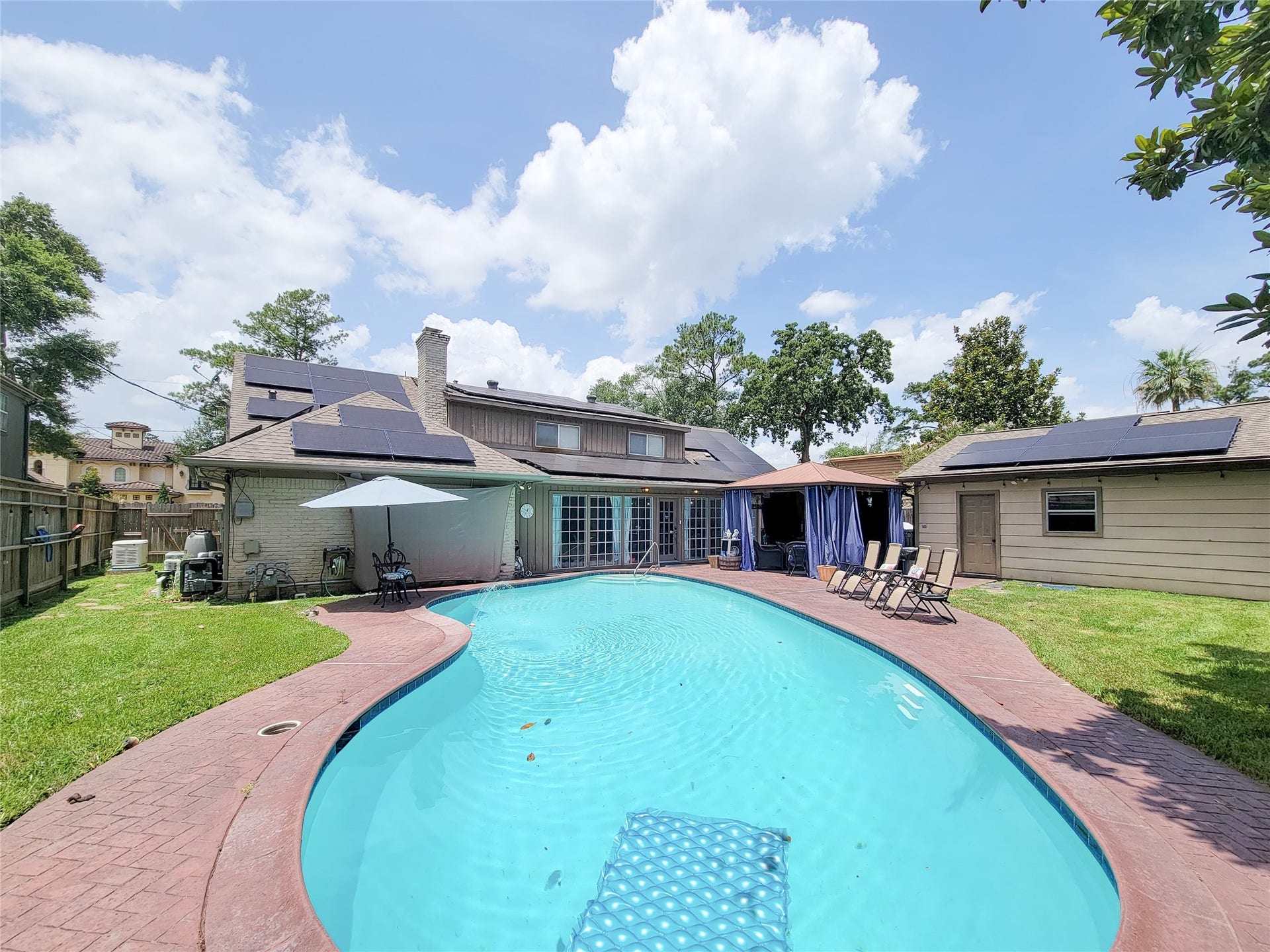 6235 Rolling Water Drive, Houston, TX 77069 This backyard features a spacious swimming pool with a curved design, surrounded by a paved deck. There's a cozy seating area with an umbrella and a shaded patio with curtains for privacy. Solar panels are installed on the roof, and the area is fenced for added security. Lush greenery completes the serene setting.