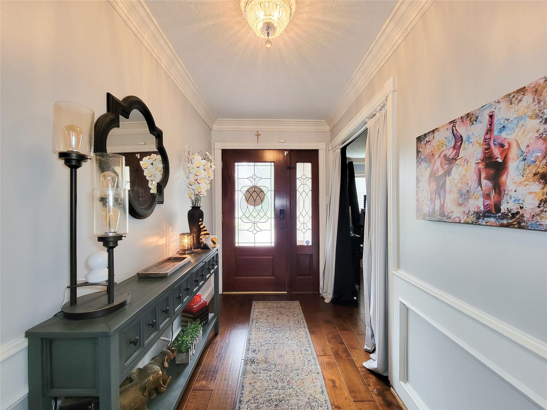 6235 Rolling Water Drive, Houston, TX 77069 This welcoming entryway features a wooden front door with decorative glass panels, elegant wainscoting, and hardwood flooring. A stylish console table with mirror and art accents adds charm, complemented by soft lighting from a ceiling fixture.