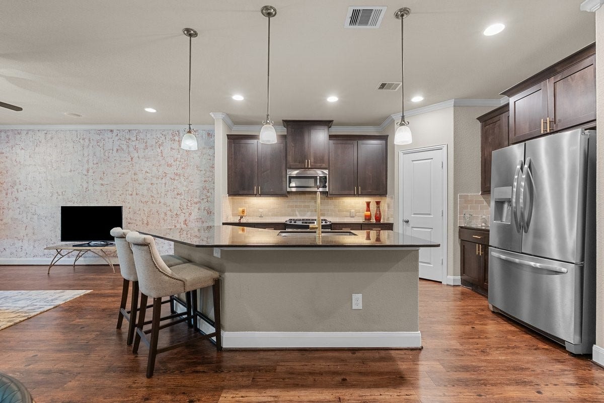 416 Via Regatta Drive, Webster, TX 77598 The kitchen shines with a curved breakfast bar, dark cabinetry, stainless steel appliances, and modern pendant lighting. Perfect for effortless entertaining and everyday luxury!