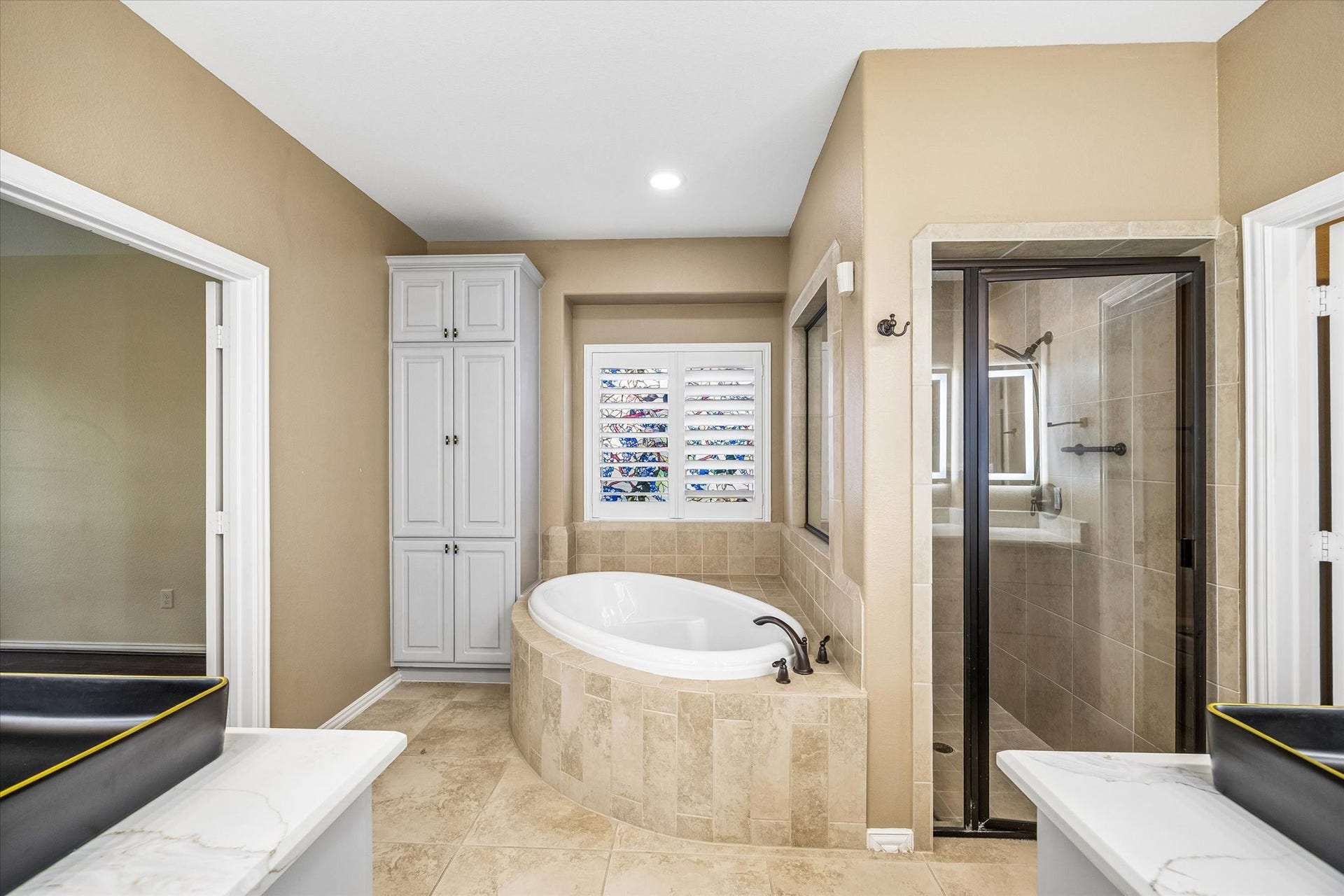 The primary en-suite bath offers separate vanities with vessel sinks, a soaking tub beneath a shuttered window and a glass-enclosed shower.  A tall linen cabinet provides ample storage.
