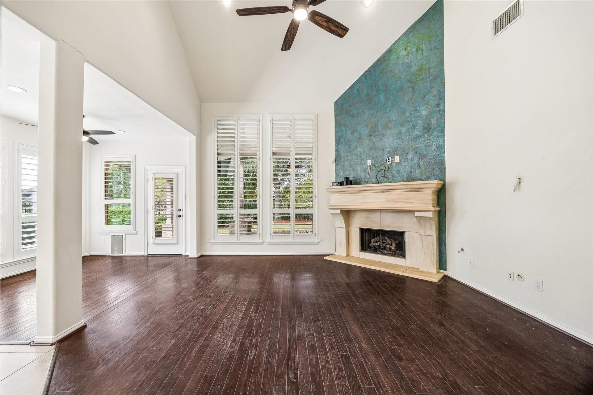 Family room with soaring ceiling, tall windows with plantation shutters and wood flooring.