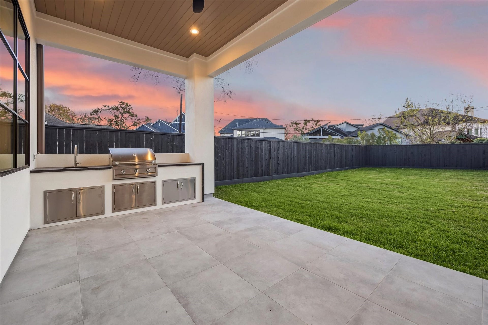 Covered patio designed for effortless outdoor living, featuring a built in grill, sink, and storage, all tucked beneath a wood paneled ceiling with recessed lighting. Spacious layout opens to a wide, grassy yard framed by a privacy fence, offering plenty of room to relax, entertain, or plan for a future pool while enjoying sunset views.**Previous build by Arch Angelo**
