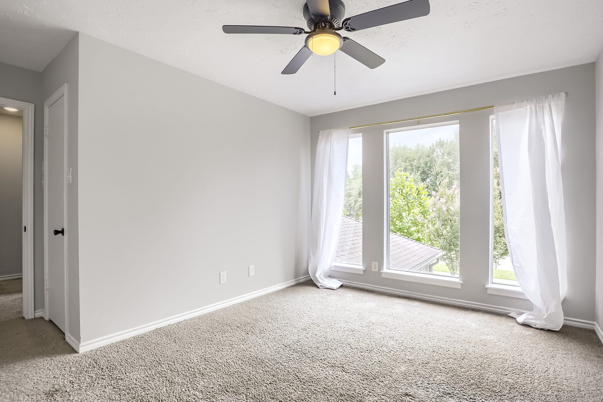 16505 Cornwall Street, Jersey Village, TX 77040 Bright bedroom with large windows, light gray walls, and soft carpet. Features a ceiling fan and white curtains, creating a cozy and airy atmosphere.