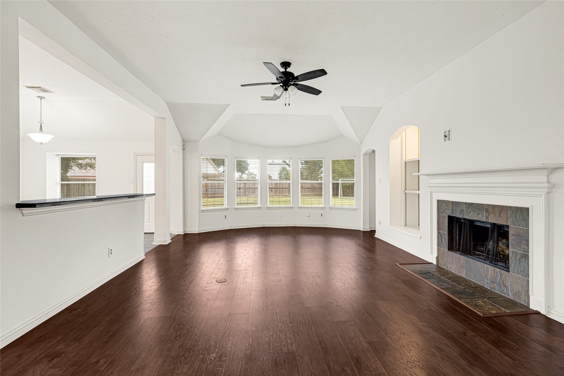 7102 Hollow Field Lane, Cypress, TX 77433 Expansive living room showcasing rich wood-look flooring, a cozy fireplace with natural stone tile accents, and a distinctive multi-window bay bump-out that bathes the space in natural light.