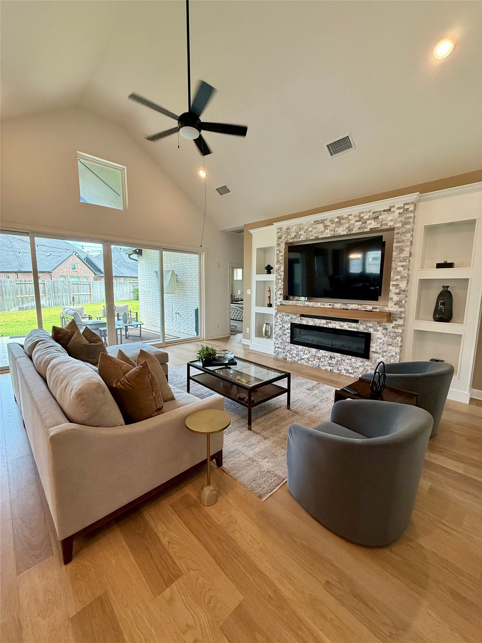 9911 Deep Water Drive, Missouri City, TX 77459 Spacious living room with high ceilings, elegant wood flooring, and custom designed modern stone accent wall featuring a built-in fireplace and TV. Large windows provide ample natural light and offer views of the backyard.