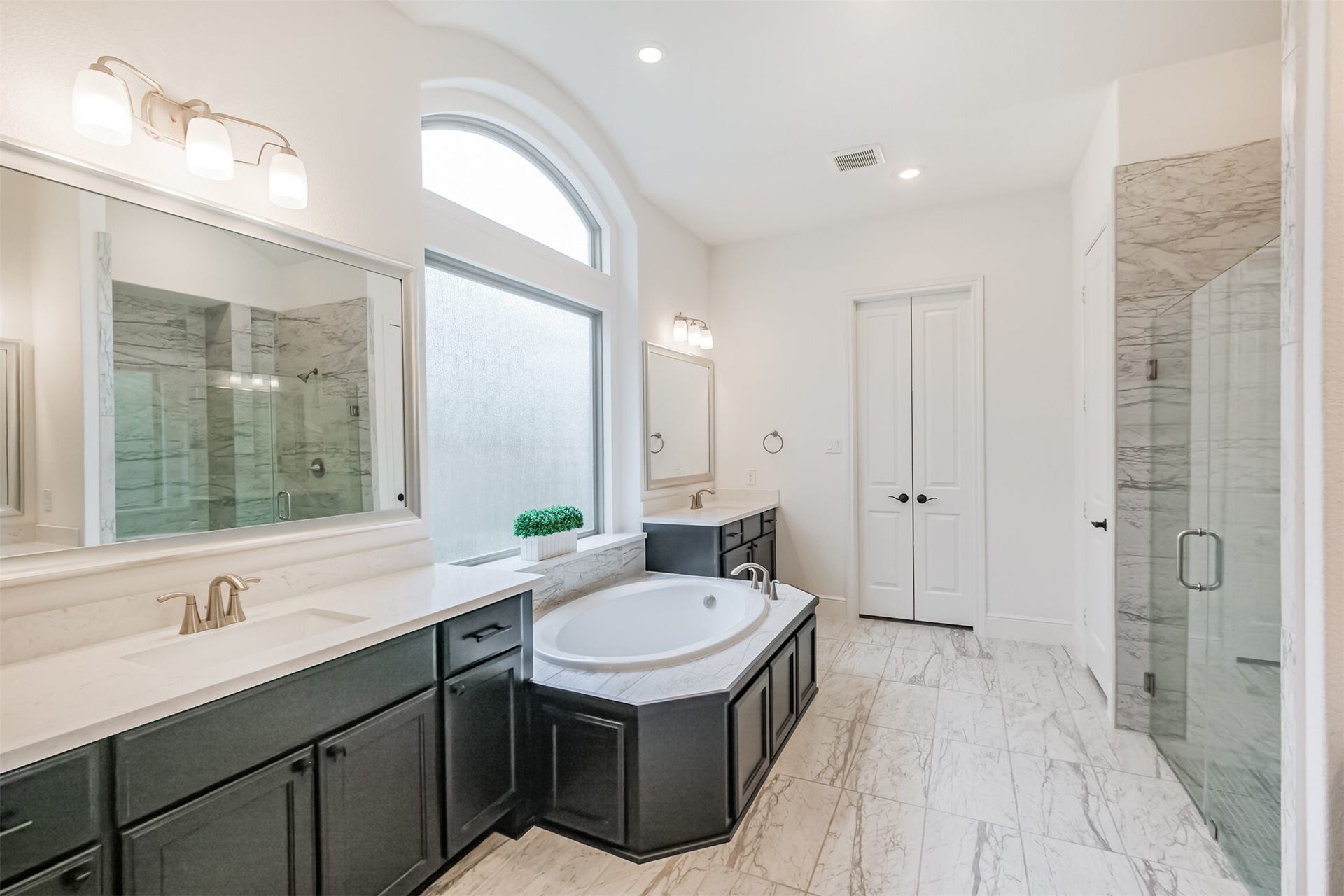 13611 Saddlebred Springs Lane, Cypress, TX 77429 Luxurious primary bathroom designed as a serene retreat, featuring a spacious soaking tub, dual vanities, elegant marble tile flooring, and a glass-enclosed shower. Abundant natural light enhances the bright, spa-like atmosphere and sophisticated finishes throughout.