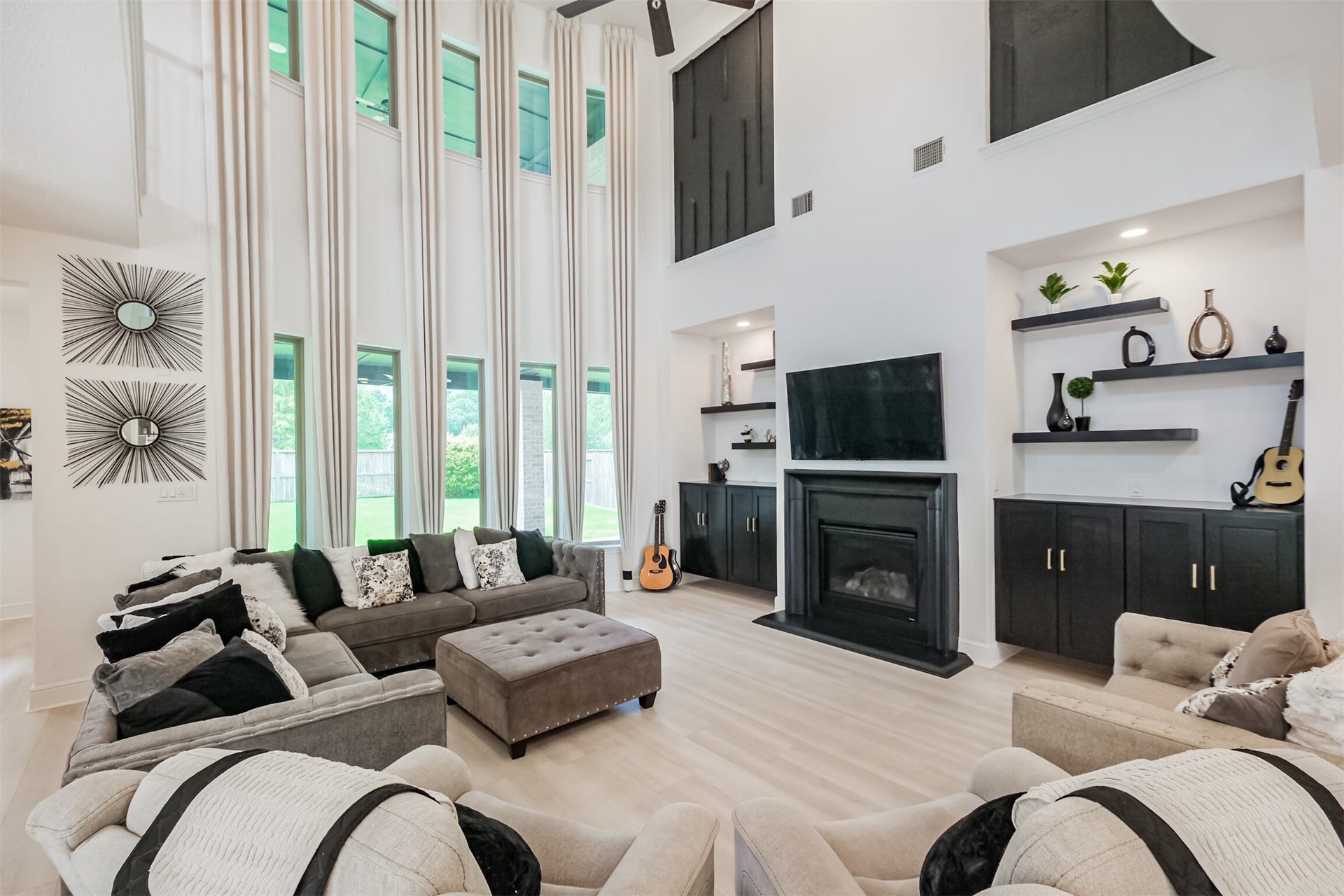 13611 Saddlebred Springs Lane, Cypress, TX 77429 Grand living room showcasing dramatic 25-foot ceilings, expansive bay windows, and abundant natural light. Custom drapery, floating cabinetry and shelving, bespoke architectural accents, and a sleek electric fireplace elevate the space, while impressive 8-foot wood-look plank flooring adds warmth and sophistication throughout.